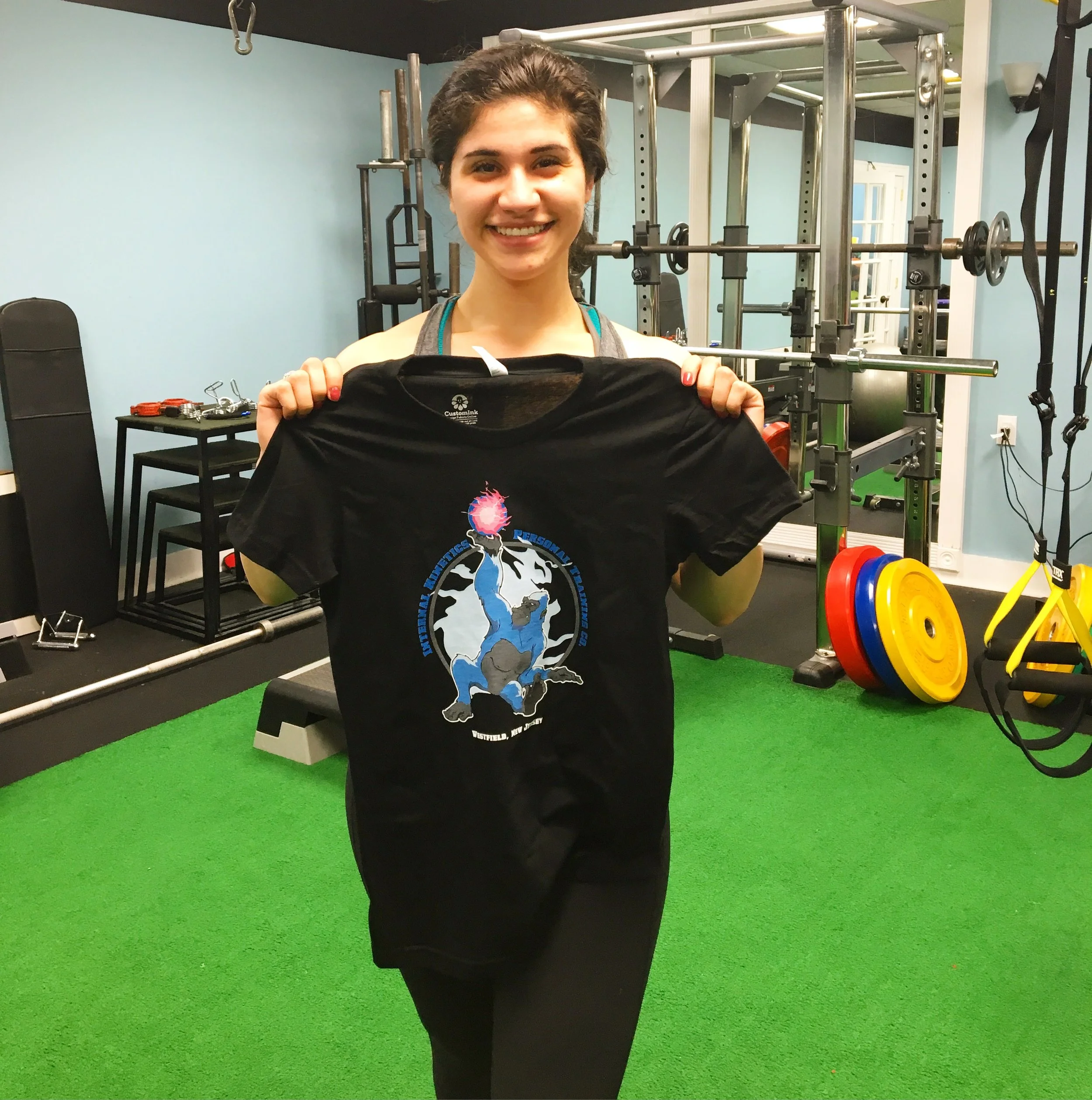 A young woman smiling and holding a black shirt inside a gym with weightlifting equipment, weights, and a green rubber floor.