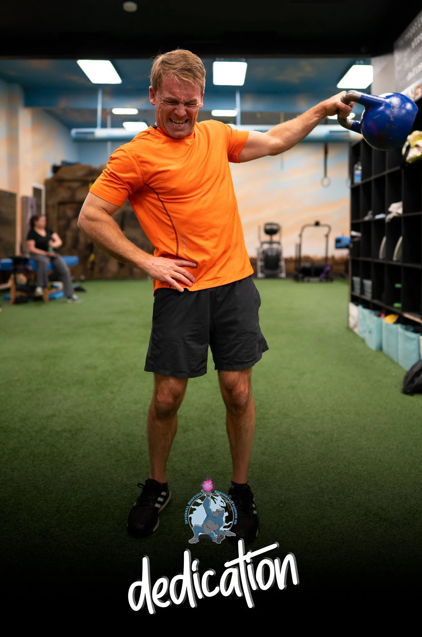 A man in an orange shirt and black shorts at a gym, holding a kettlebell while grimacing. In the background, a woman is sitting on a bench. The floor is green turf, and there are gym equipment and lockers visible. There is a logo with a brain and glo