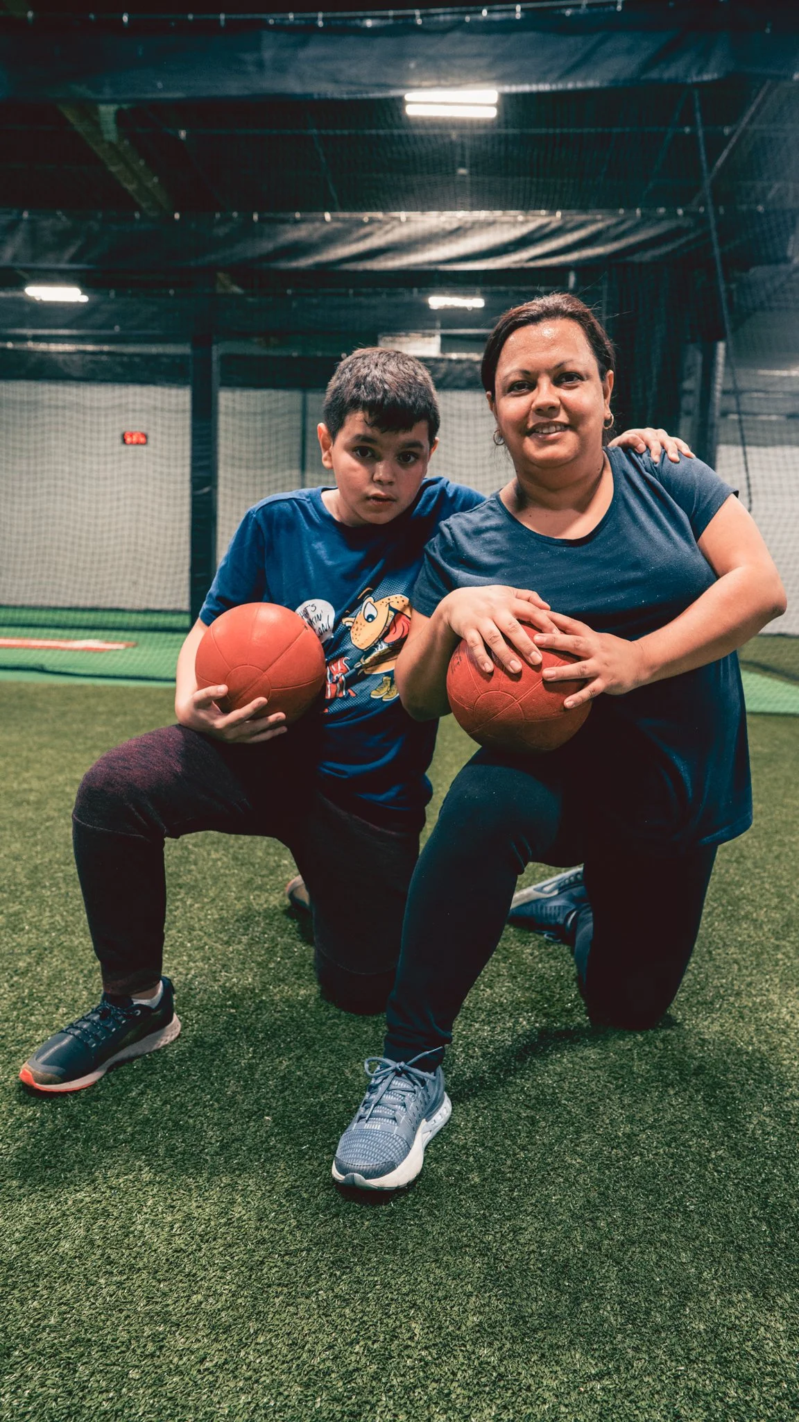 A woman and boy kneeling on an indoor sports court, holding basketballs and smiling at the camera.
