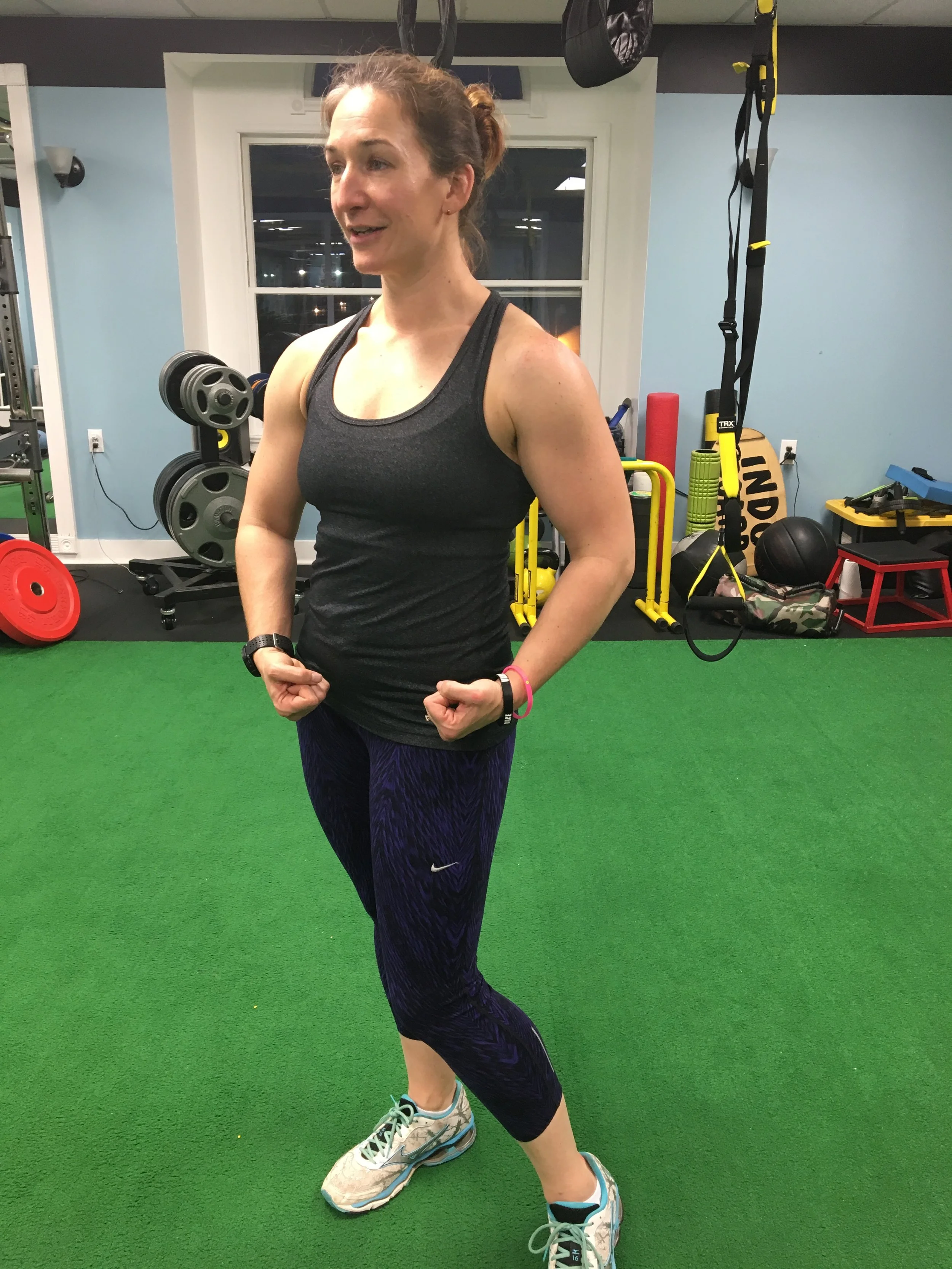 A woman in workout attire flexing her biceps in a gym with various exercise equipment and a window in the background.