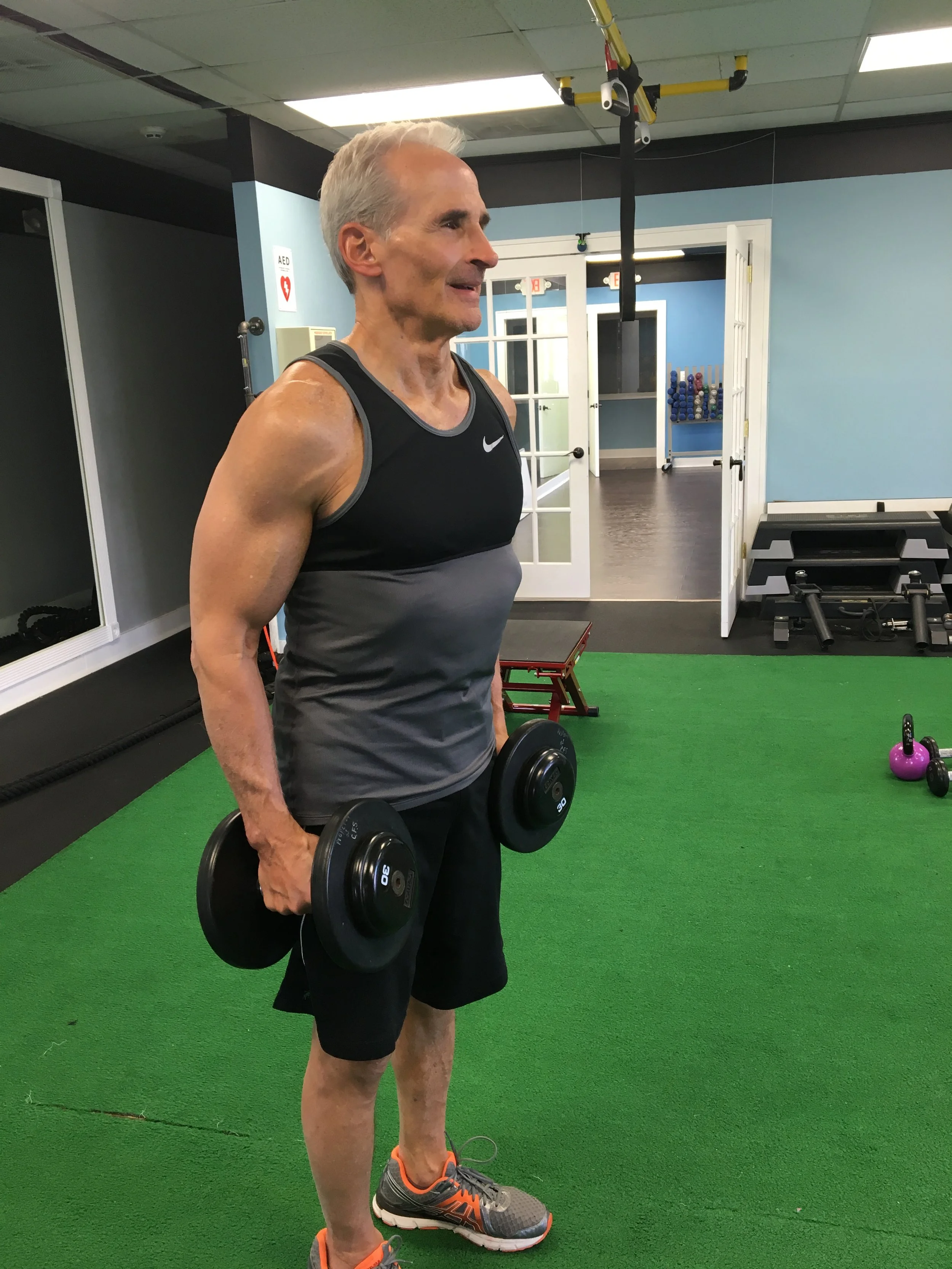 An older man with gray hair wearing a black and gray Nike tank top and black shorts, standing on a green turf floor, holding two dumbbells, inside a gym with blue walls, and various exercise equipment in the background.