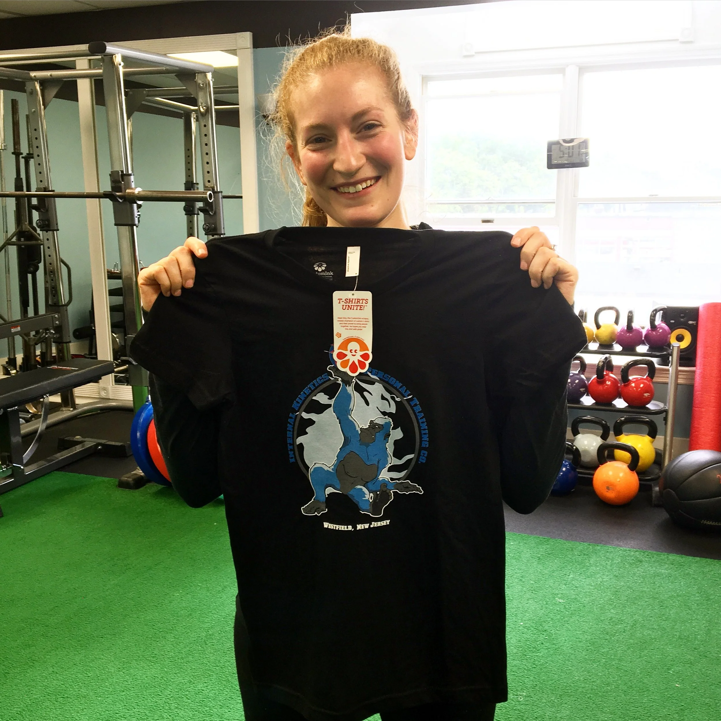 A woman smiling in a gym holding a black T-shirt with a tag that reads 'T-SHIRTS UNITE!'. There are kettlebells and gym equipment in the background.