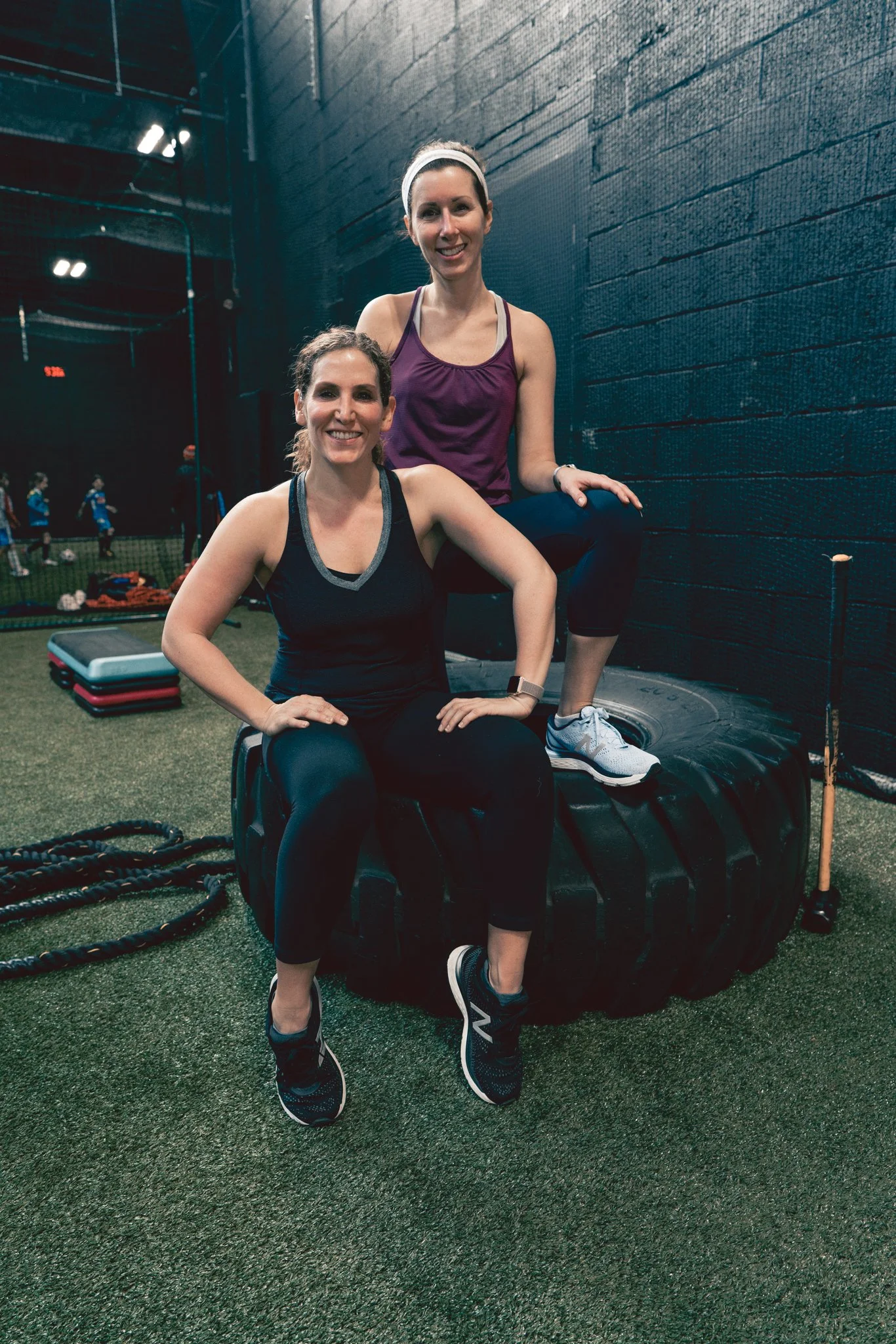 Two women in workout attire in a gym, one sitting on a large tire and the other standing behind her with one foot on the tire, smiling at the camera.