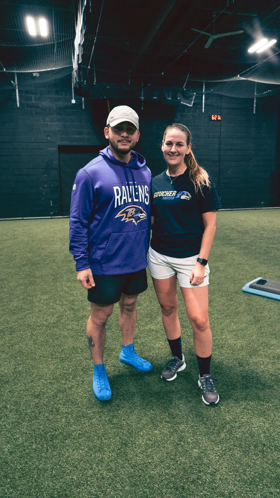 A man and woman standing together on an indoor sports field, smiling at the camera, with the man wearing a purple Baltimore Ravens hoodie and the woman wearing a black soccer Goucher shirt.