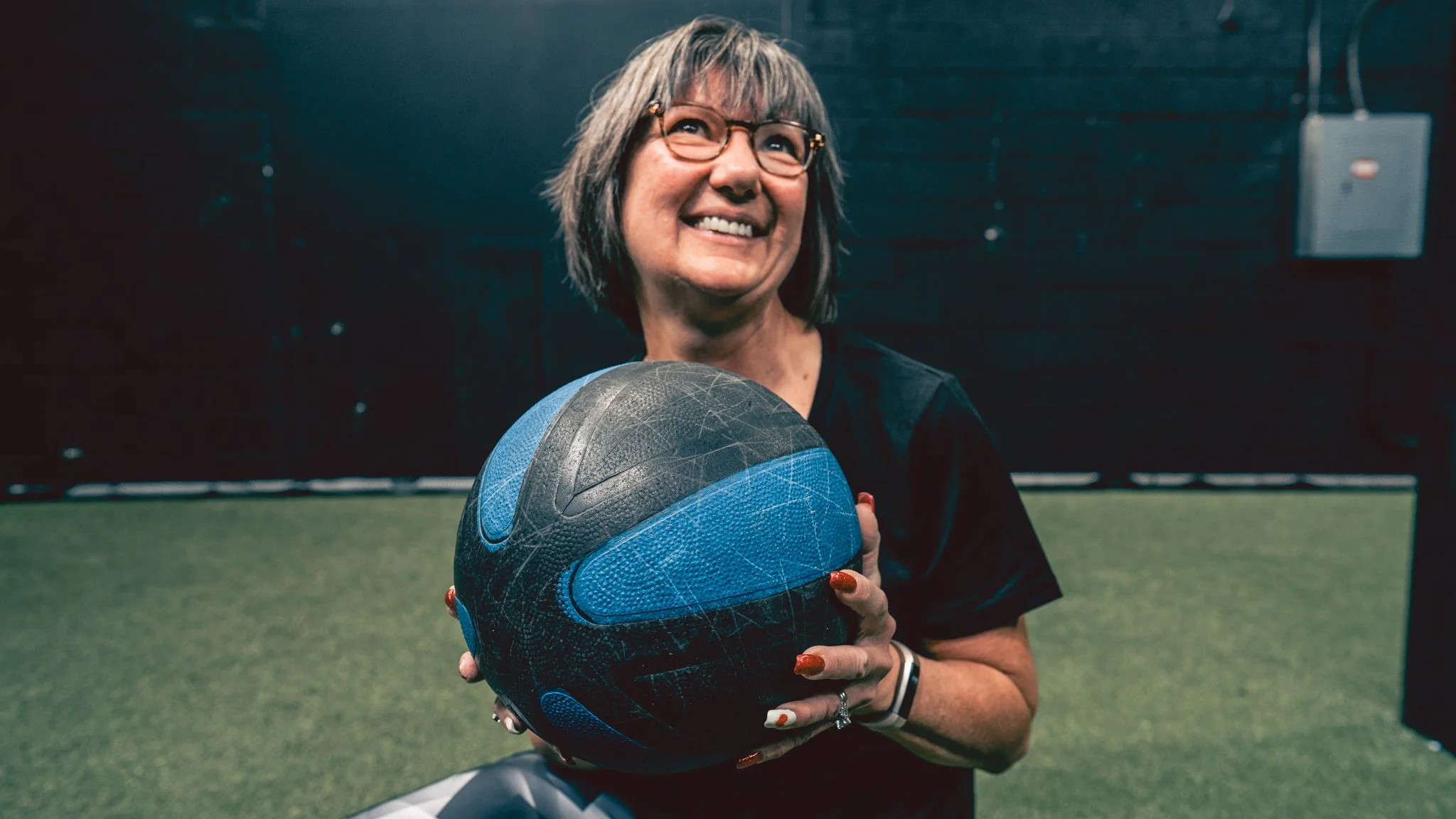A woman with glasses and gray hair smiling and holding a blue and black medicine ball indoors on a green turf field.