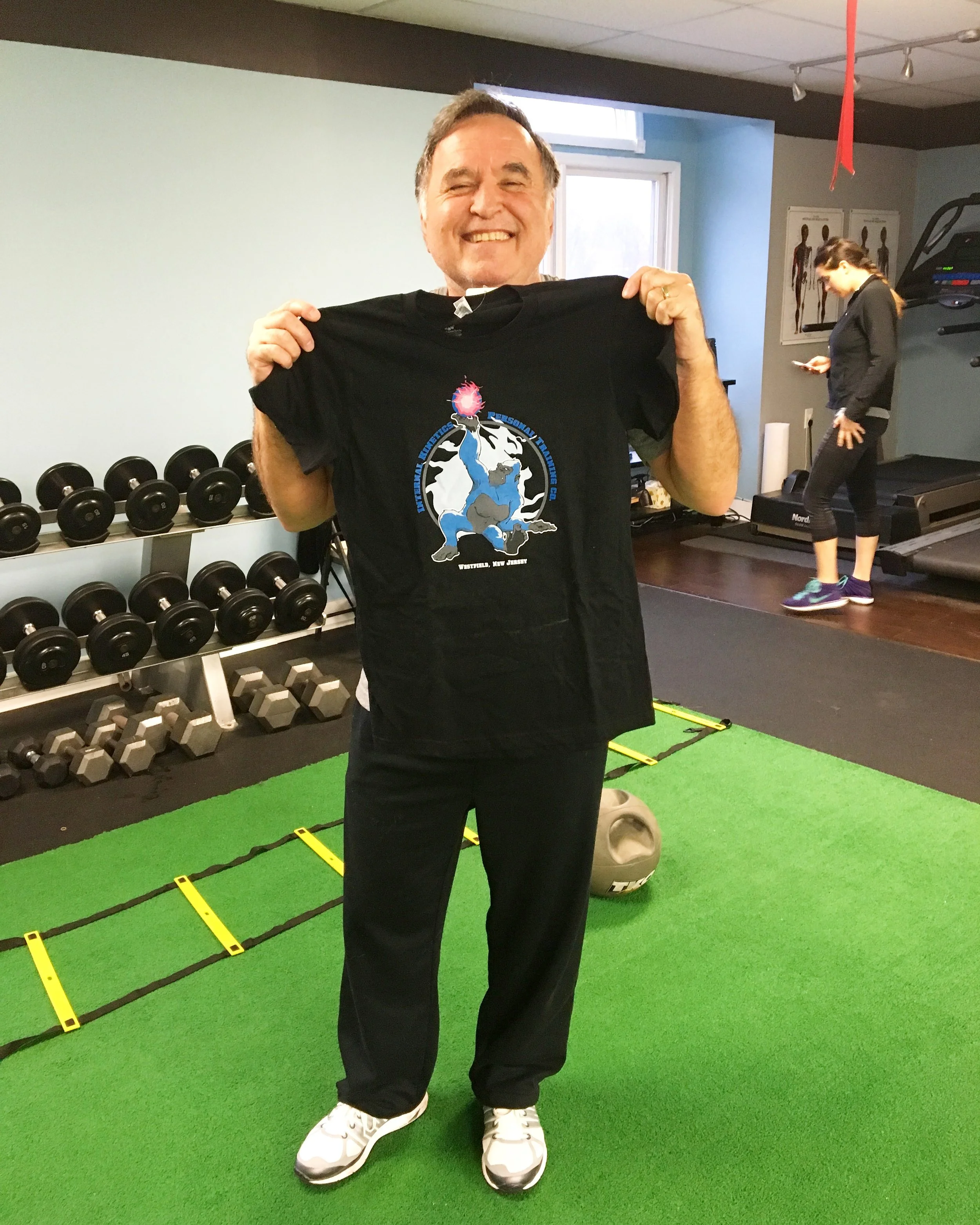 A smiling man holding a black t-shirt with a cartoon dinosaur graphic in a gym or fitness studio. The background shows dumbbells, a treadmill, and a person working out.
