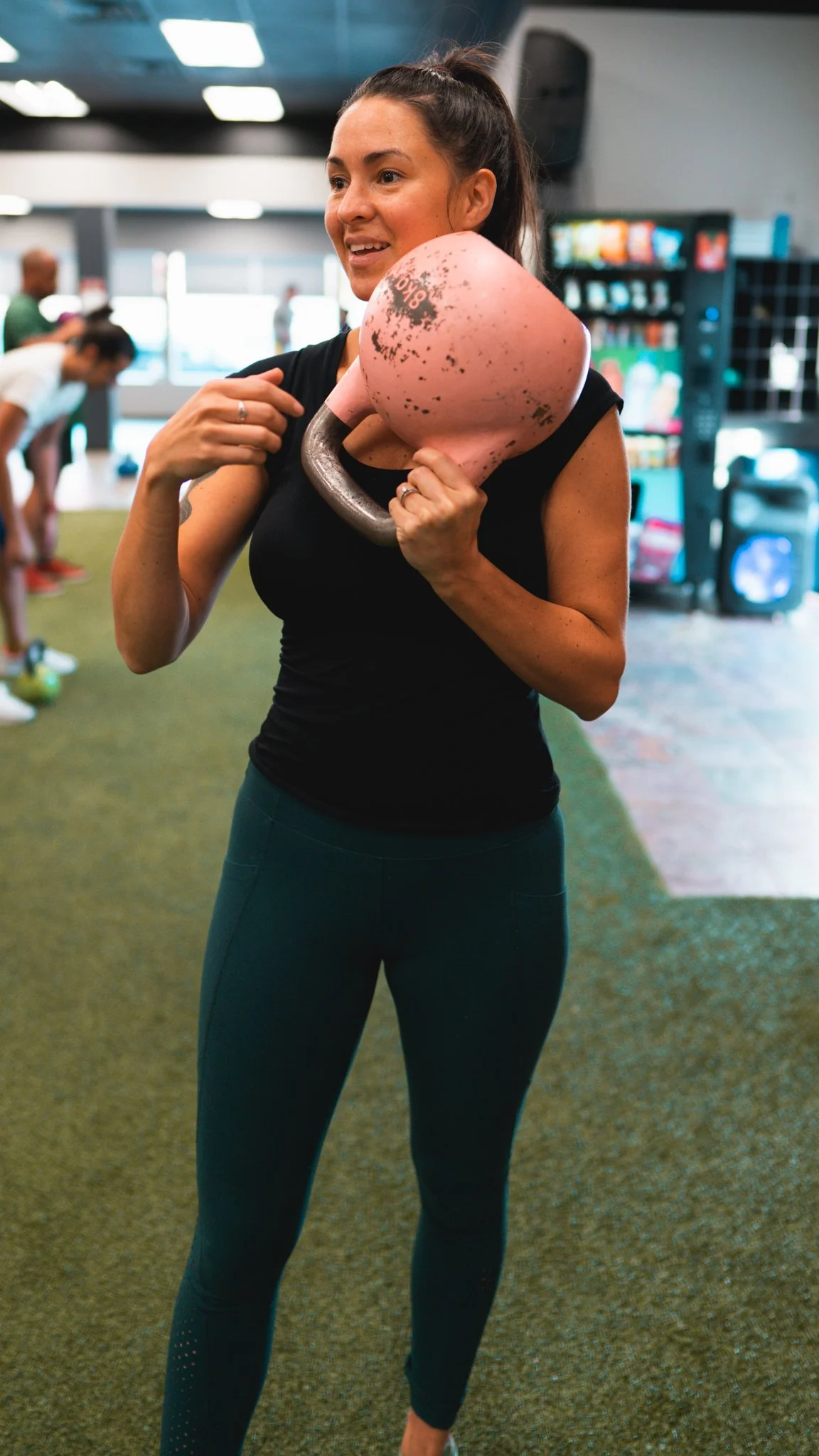 A woman with a ponytail holding a pink kettlebell in a gym.