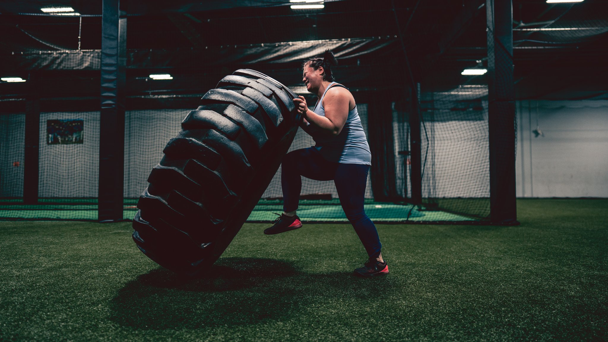 A woman in workout attire, including a gray tank top and dark leggings, is lifting a large tire in an indoor gym with green turf flooring. The gym has netted walls and dim lighting.