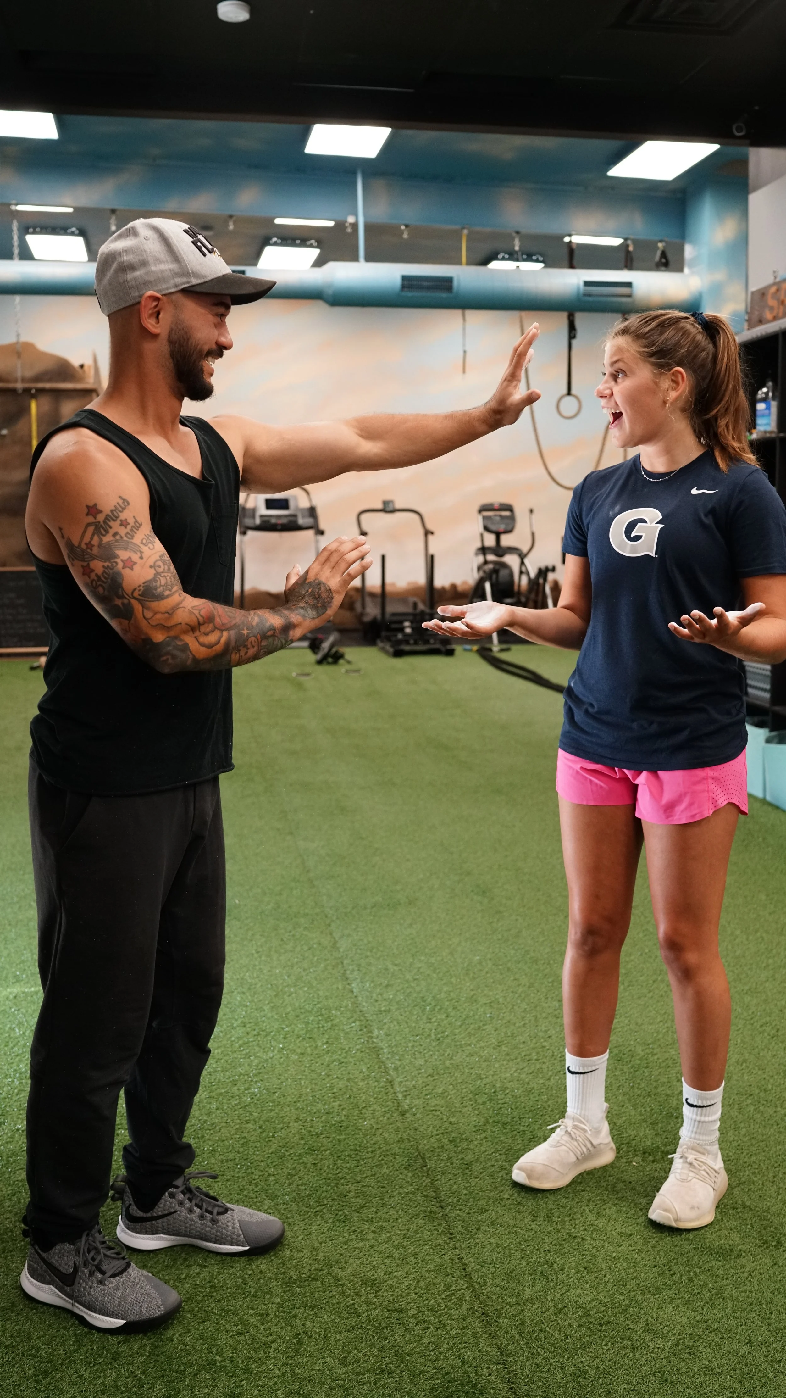 A man and a young woman in a gym. The man is giving a high five to the woman. The man has a beard, tattoos, and is wearing a gray cap, black tank top, black pants, and gray sneakers. The woman has long hair tied back, and is wearing a navy blue t-shi