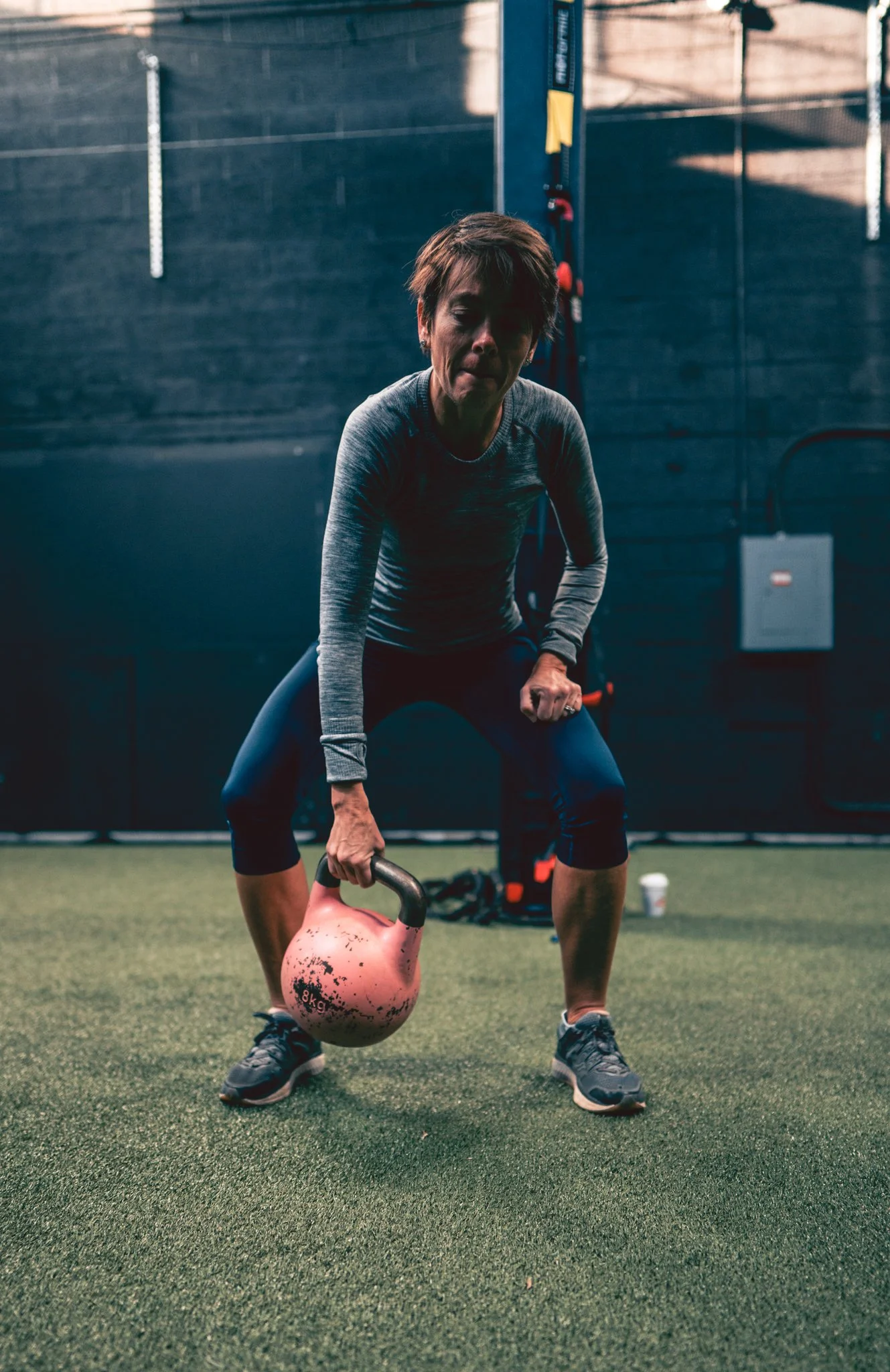 A woman in workout clothes performs a kettlebell exercise indoors on a green turf floor, with gym equipment and a wall in the background.