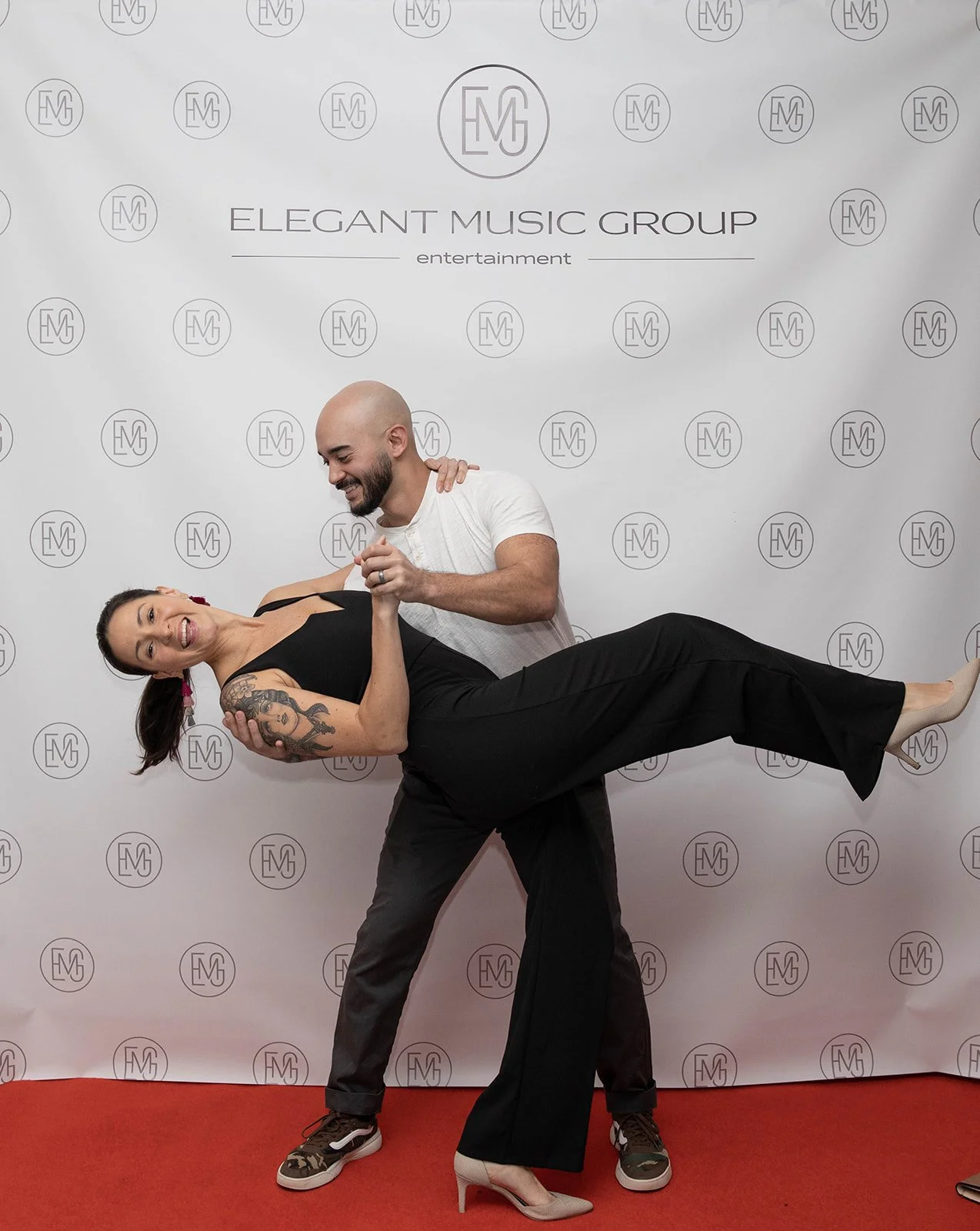 A man and woman dancing at an event with a backdrop reading 'Elegant Music Group entertainment.' The man is holding the woman in a dance pose, smiling, with the woman smiling and leaning back. The woman has a tattoo on her arm, wears a black outfit, and high heels. The man is dressed casually in a white t-shirt, dark pants, and sneakers.