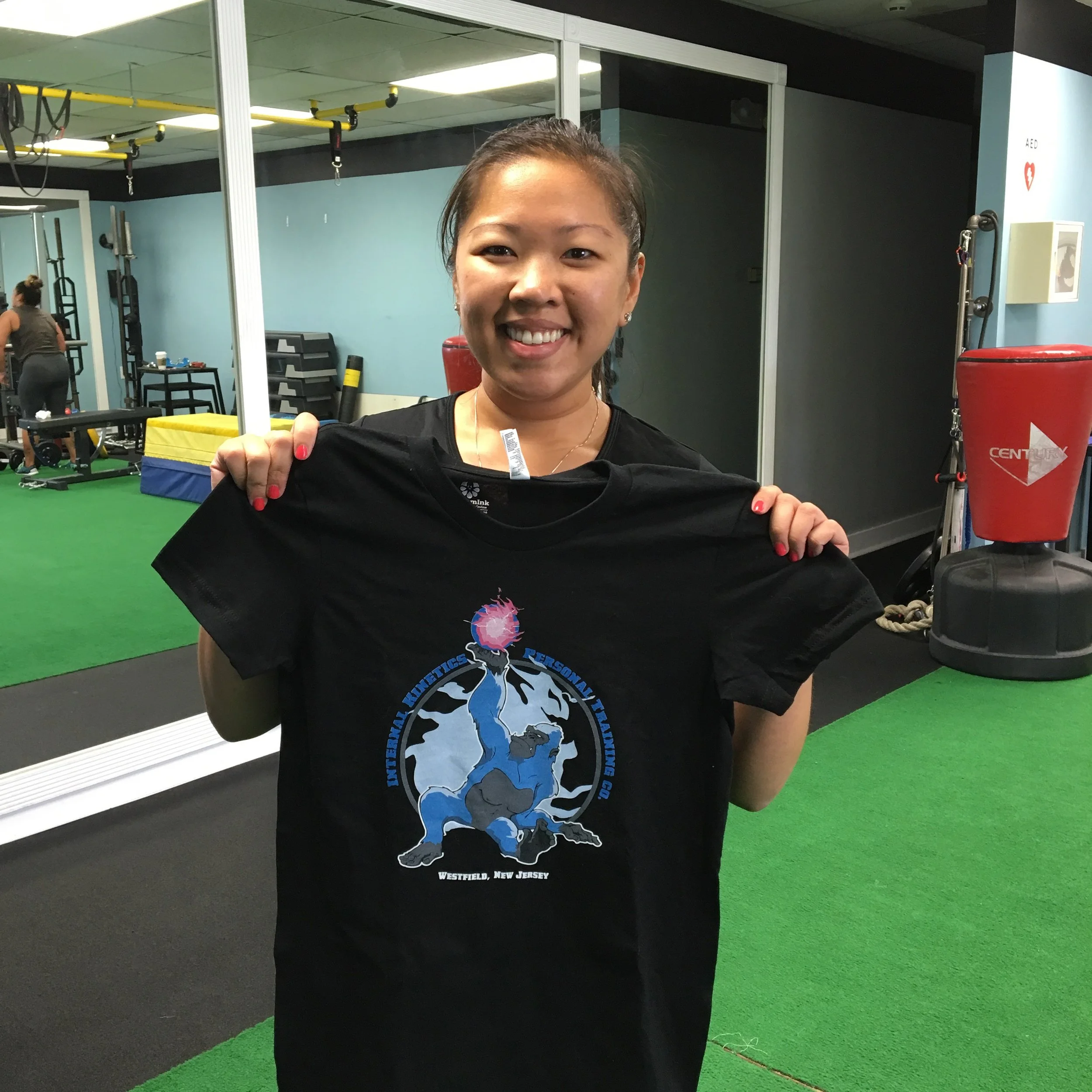 A smiling woman holding a black t-shirt in a gym. The t-shirt has a graphic of a lion and text. The gym has blue walls, gym equipment, and a green and black floor.