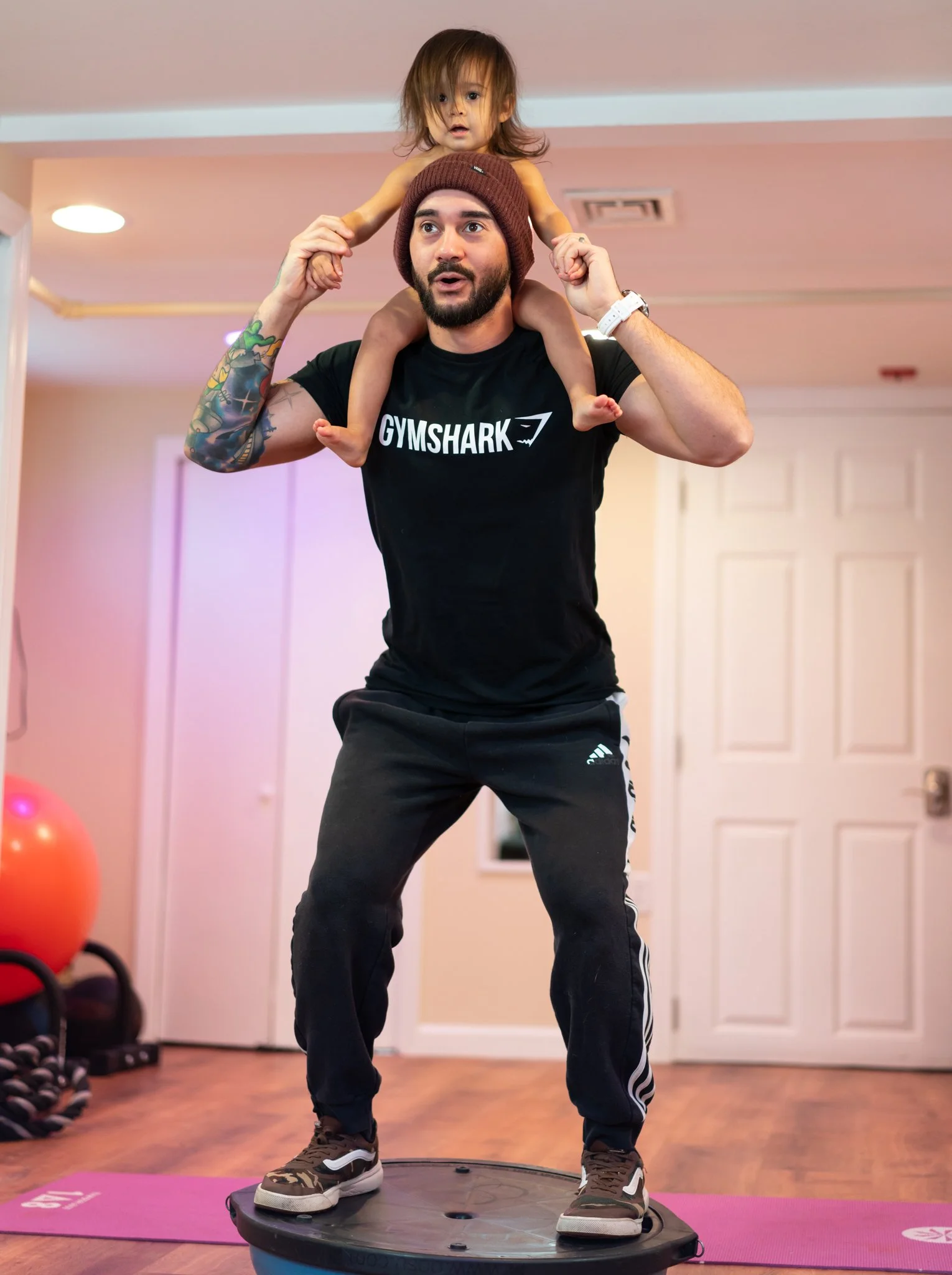 A man with a beard, wearing a brown beanie and a black Gymshark t-shirt, is standing on a black balance trainer. A young girl with brown hair, wearing a yellow tank top, is sitting on his shoulders. The man appears to be exercising or balancing, and the girl is looking forward with about shoulder length hair. They are indoors in a room with pink walls, wooden floors, and exercise equipment like dumbbells and a large red exercise ball visible in the background.