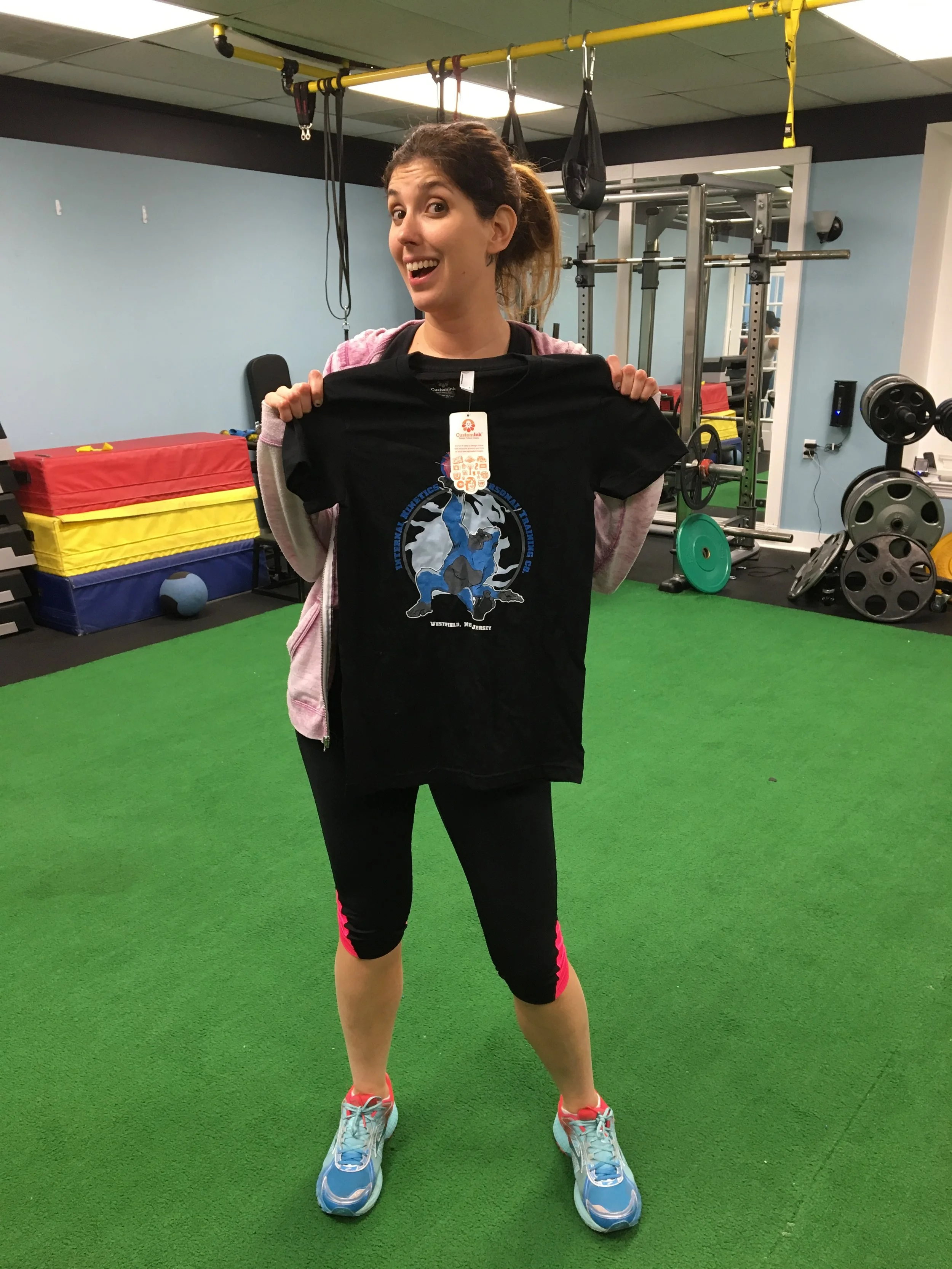 A woman standing on a green gym floor, holding a black t-shirt with a logo, in a fitness center with weightlifting equipment in the background.
