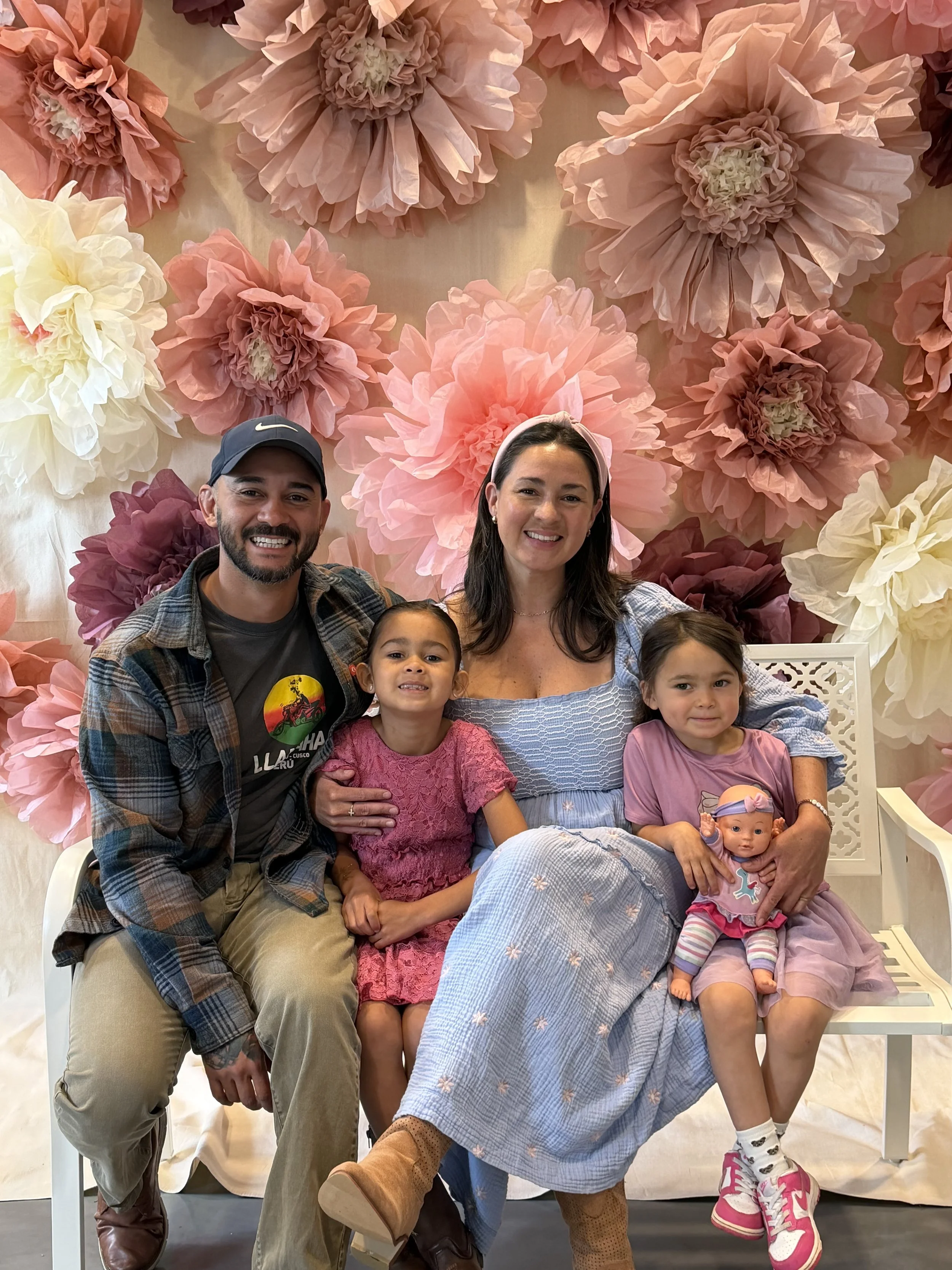 Family of five, including two children, posing for a photo in front of pink and cream paper flower backdrop. The father is wearing a baseball cap and plaid shirt, the mother is in a light blue dress with a headband, and the three girls are dressed in pink and purple, one holding a doll.