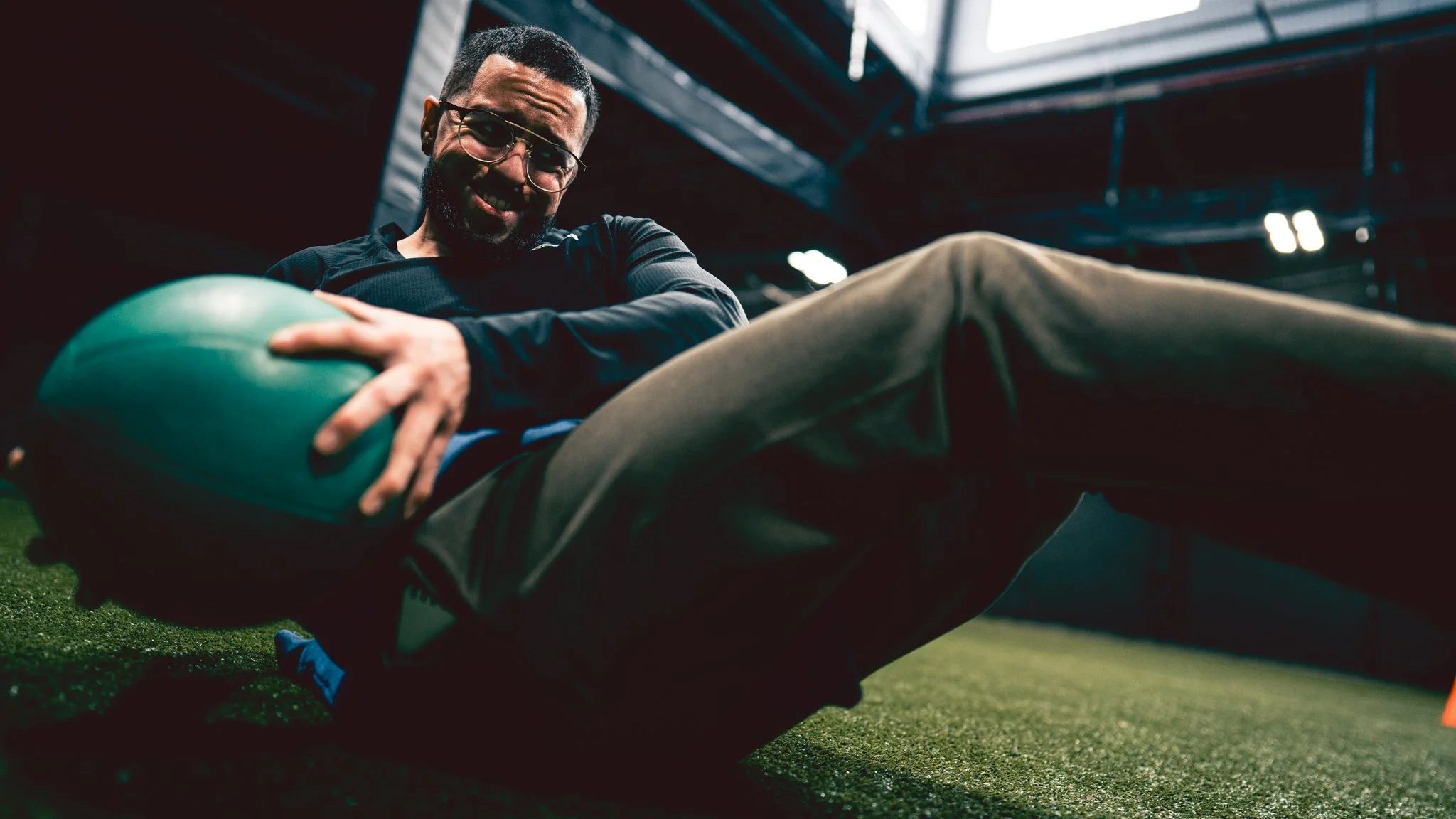 A man with glasses smiling and lying on the ground inside a sports facility, holding a green medicine ball, with gym equipment and a roof structure visible in the background.