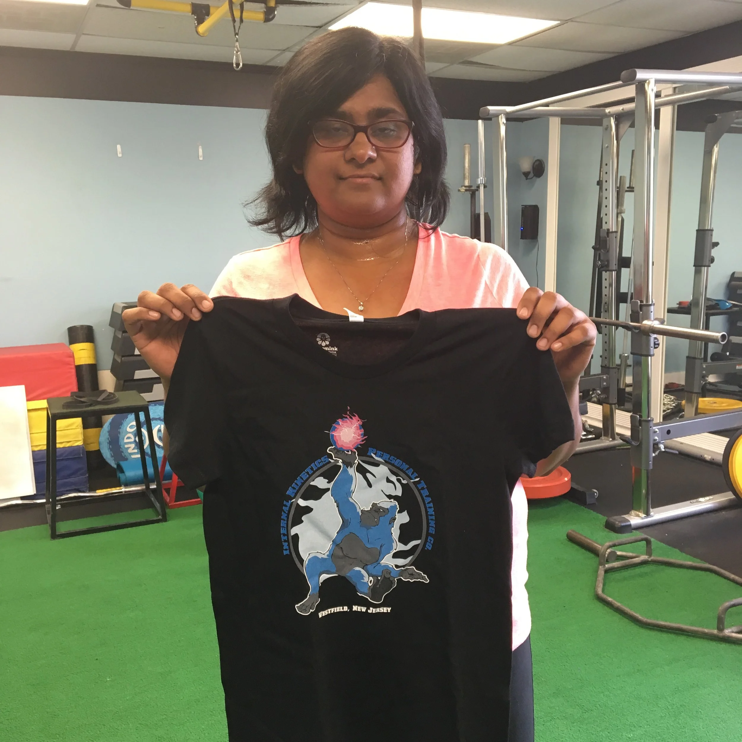 A woman with dark hair, glasses, and a pink shirt holding a black T-shirt with a logo and text, standing in a gym with workout equipment in the background.