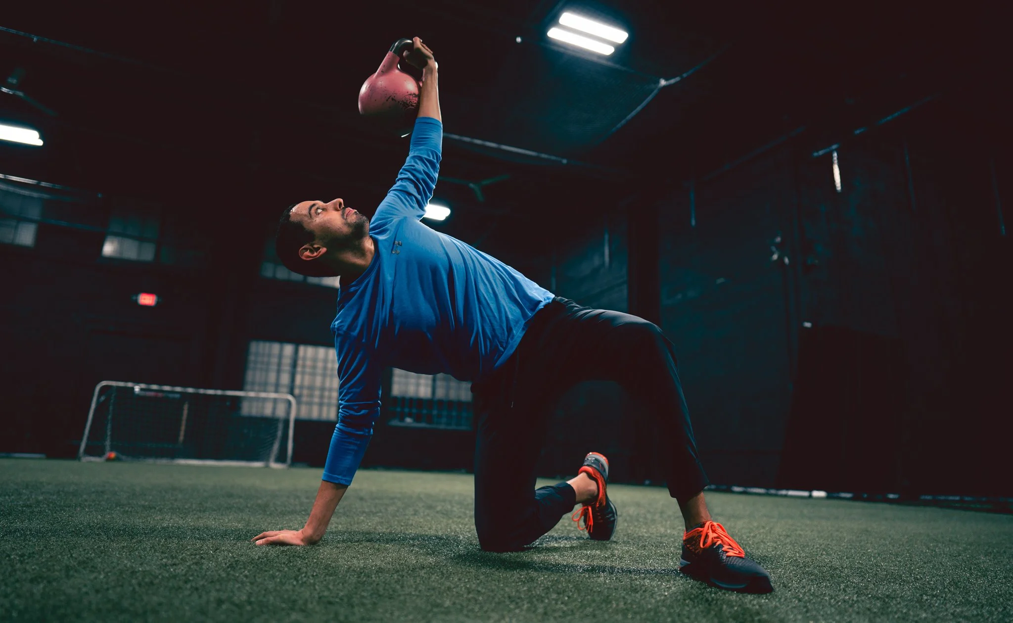 A man in a blue long-sleeve shirt and black shorts performing plow pose with a kettlebell in a gym.