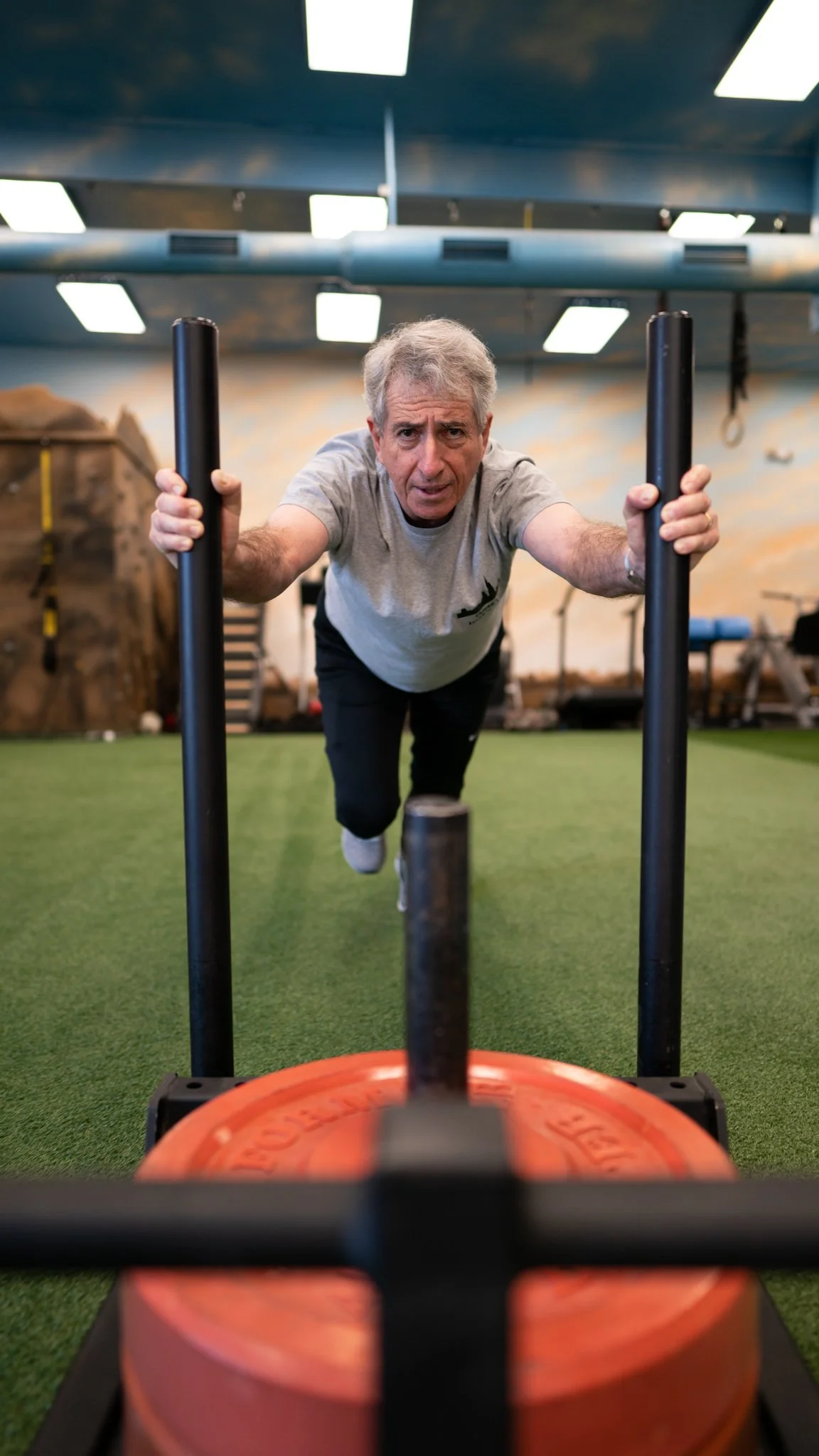 A man pushing a sled with weights at a gym, viewed from behind and close to the weights.