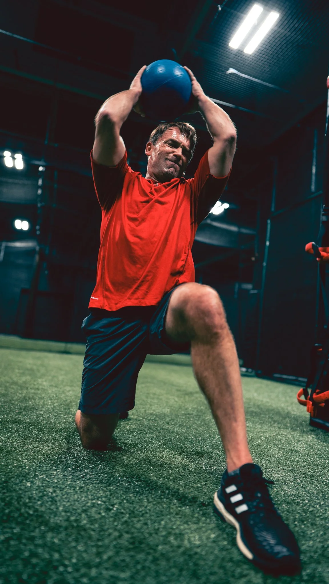 Man in red shirt and black shorts doing a knee exercise while holding a blue stability ball in a gym.