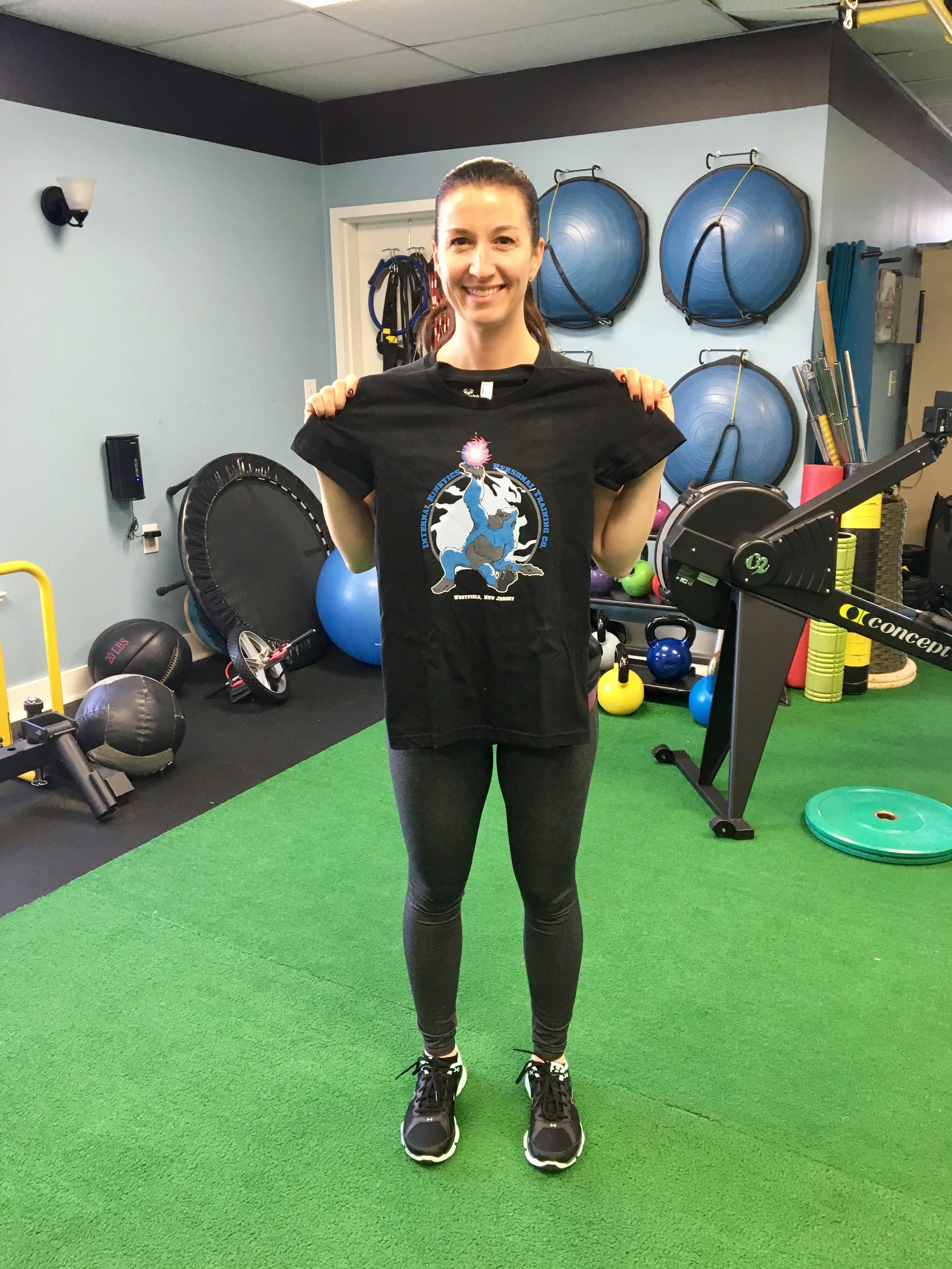 A woman smiling and holding a black T-shirt inside a gym with various workout equipment like kettlebells, medicine balls, and exercise balls in the background.