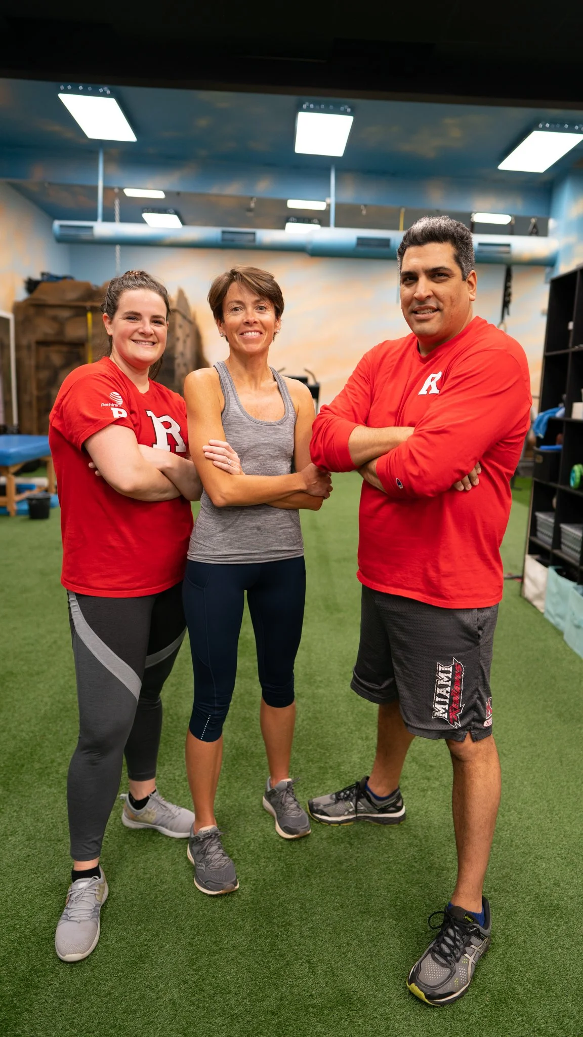 Three people standing inside a gym or fitness studio, smiling at the camera. Two women are wearing red shirts and a man is wearing a red shirt and shorts. They are standing on artificial turf, with gym equipment and mural in the background.