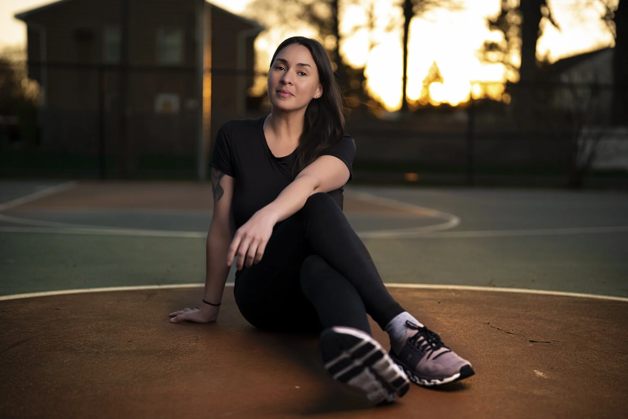 A woman sitting on a basketball court at sunset, dressed in athletic clothes, with a relaxed smile and dark hair, surrounded by trees and a building in the background.