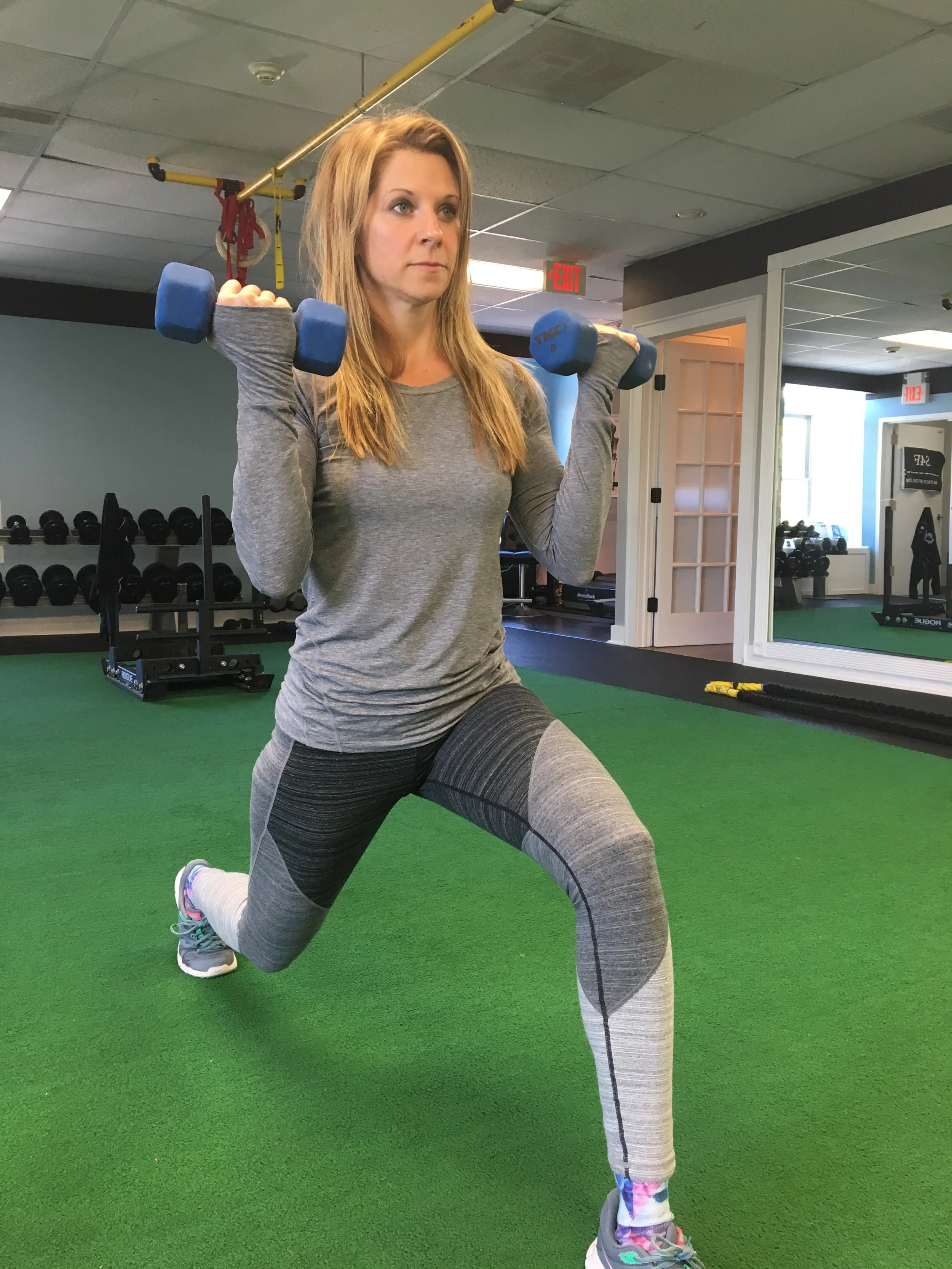 A woman in gray athletic clothing and multicolored shoes performs a lunge exercise while holding dumbbells on her shoulders inside a gym with green flooring.