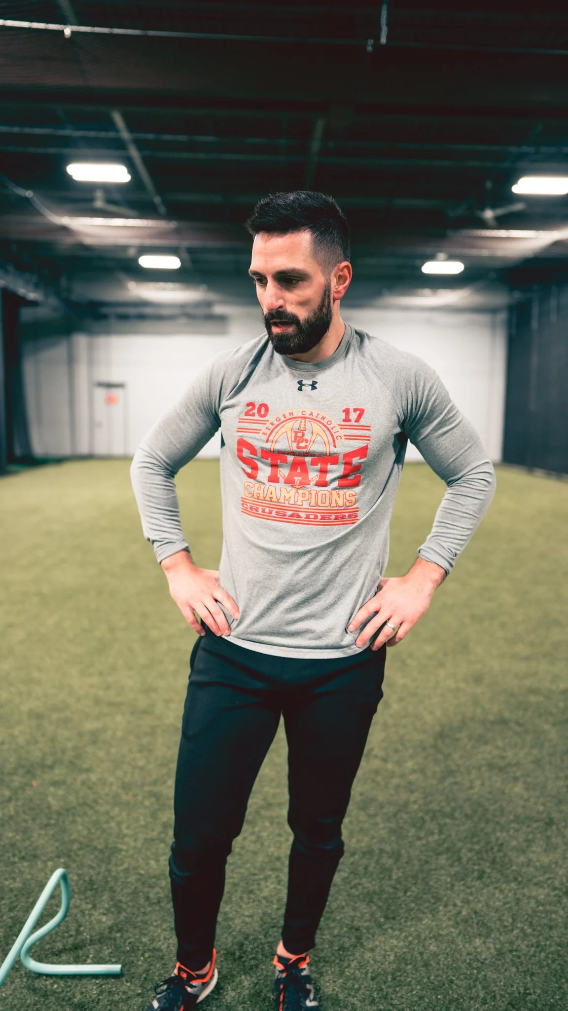 A man with a beard and short dark hair standing indoors on a green carpeted floor, wearing a grey long-sleeve athletic shirt with red lettering that reads 'STATE CHAMPIONS 2017' and black athletic pants, with his hands on his hips, looking down thoug
