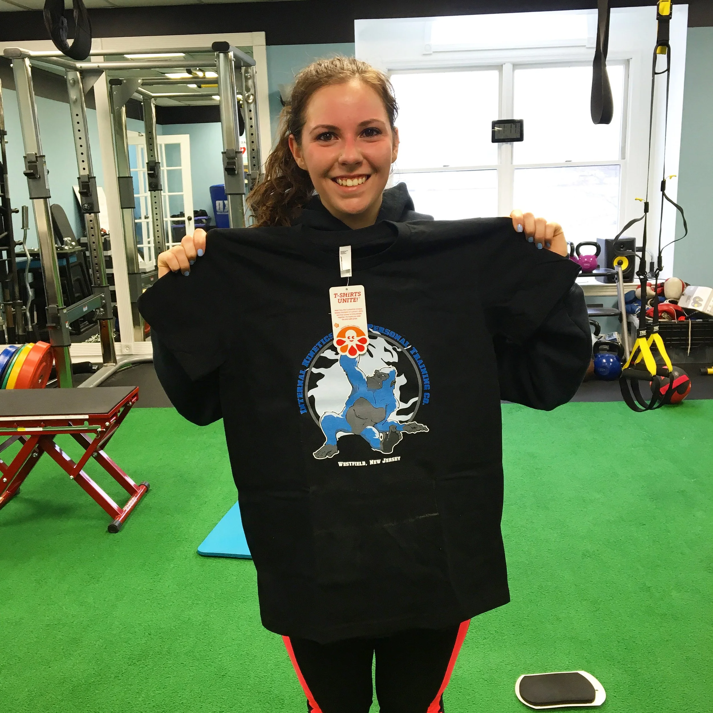 A woman smiling and holding a black t-shirt in a gym. The woman has light brown hair and is wearing a dark workout shirt with a flyer hanging around her neck. The gym has green turf flooring and workout equipment in the background, including kettlebe