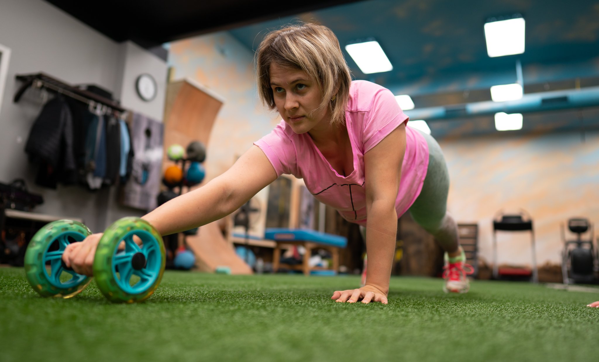 Woman in pink shirt performing a plank exercise on the floor of a fitness studio, with exercise equipment and bright lighting in the background.