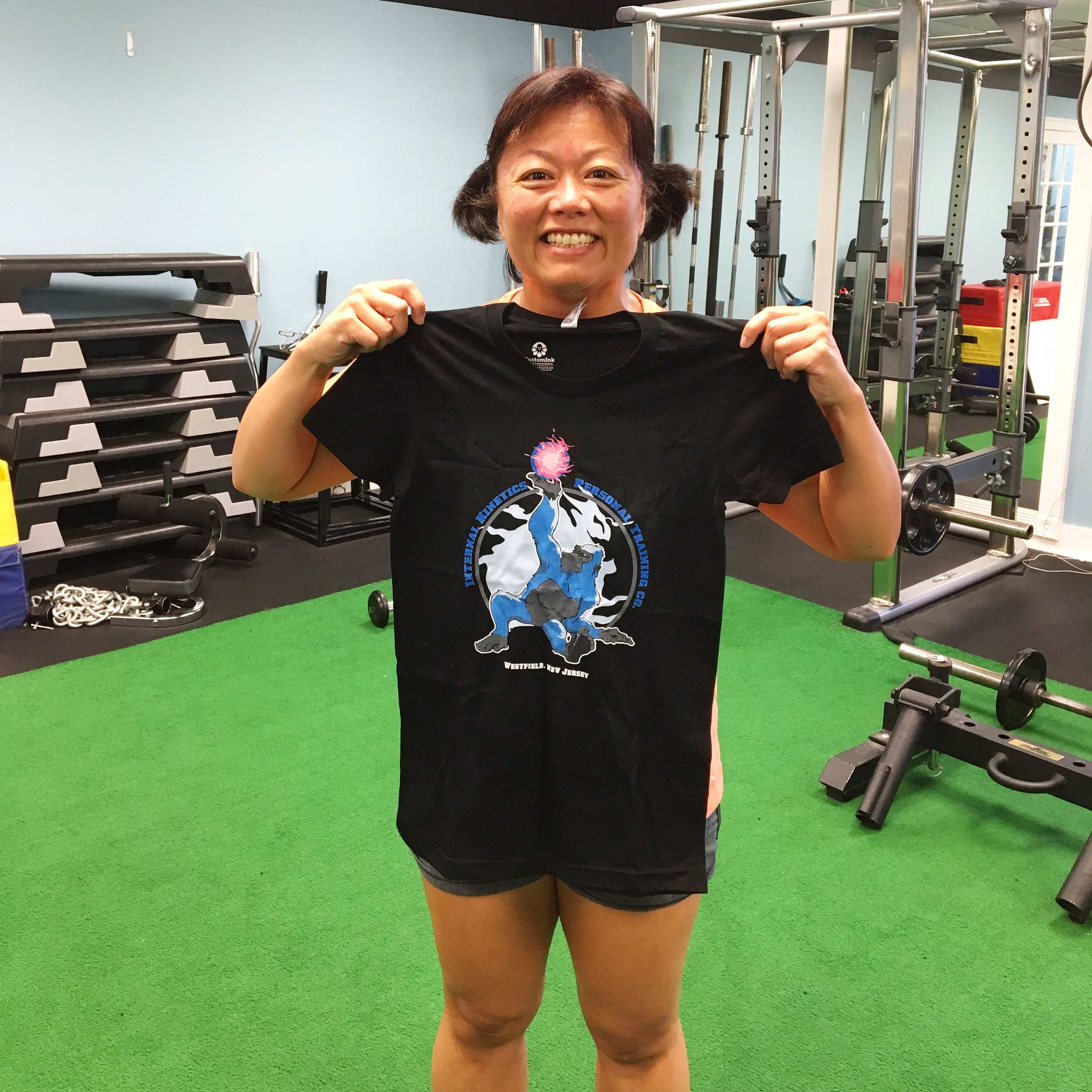 A woman smiling and holding up a black T-shirt inside a gym, with gym equipment and green turf in the background.