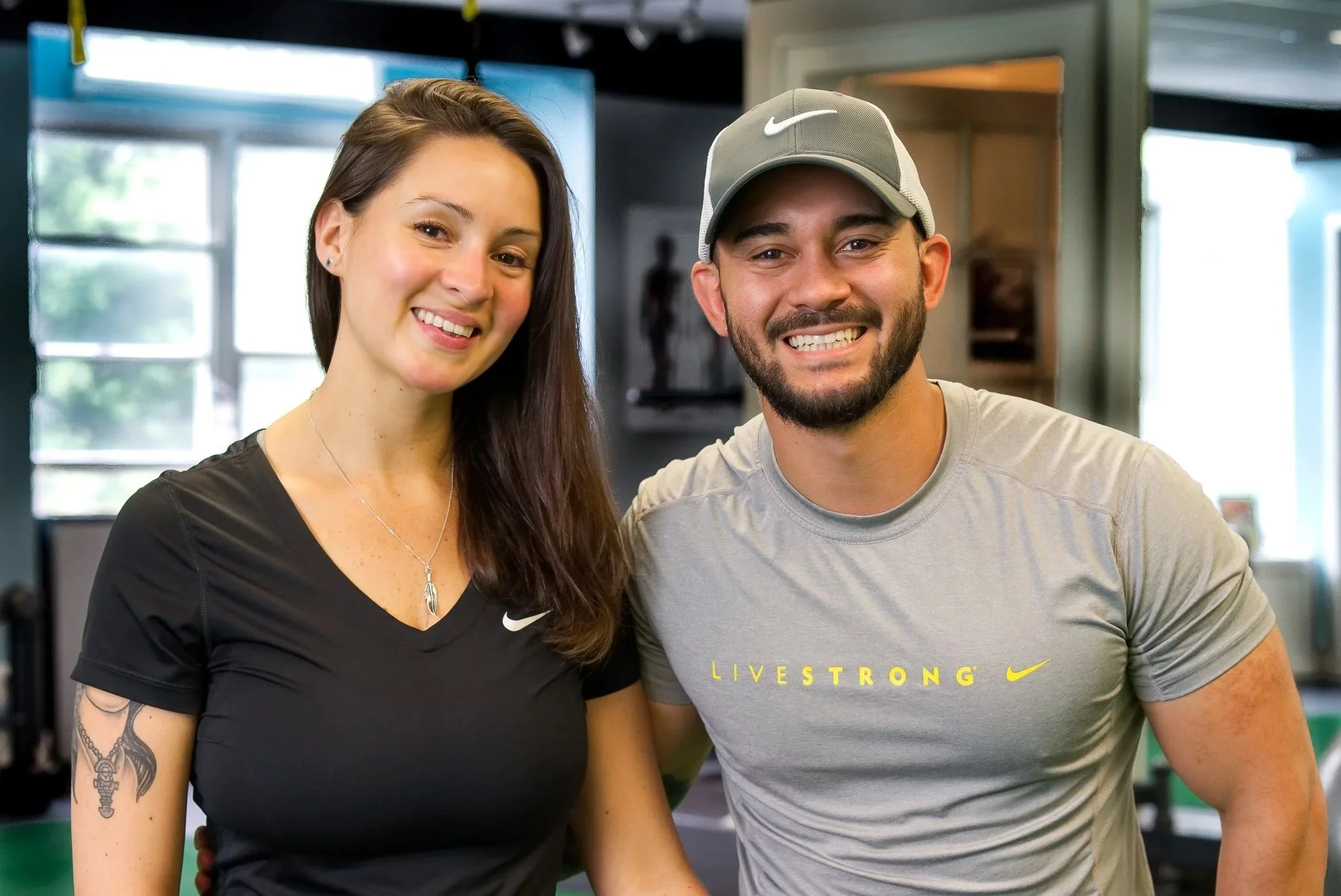 Two people smiling and posing for a photo indoors. The woman on the left has long brown hair, piercings, a tattoo on her arm, and is wearing a black Nike shirt. The man on the right has a beard, mustache, and is wearing a gray Nike hat and a gray Nike t-shirt with yellow text.