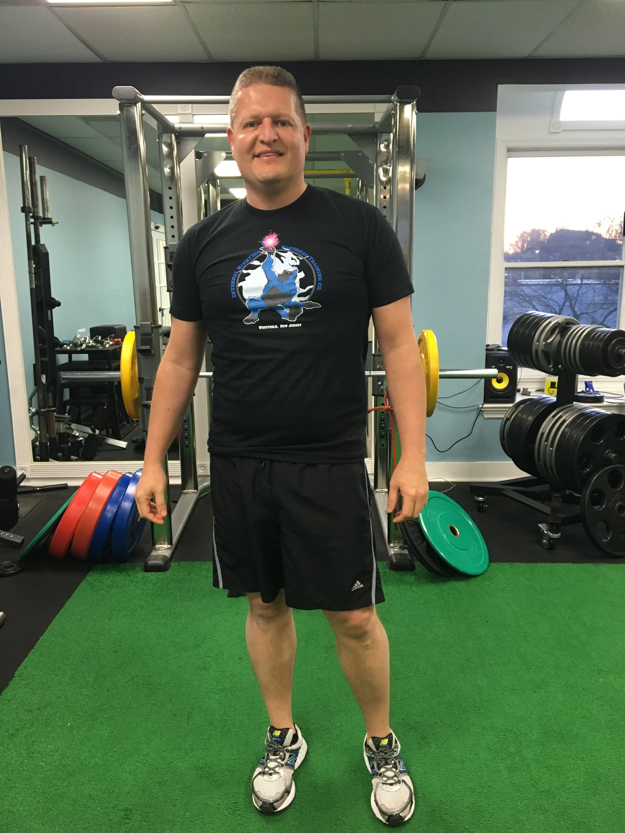 A man standing on green artificial turf in a gym, smiling, wearing black athletic shorts, a black T-shirt with a graphic of a blue dragon and the text "Westfield, New Jersey," and athletic shoes. Behind him are weightlifting equipment, including weig