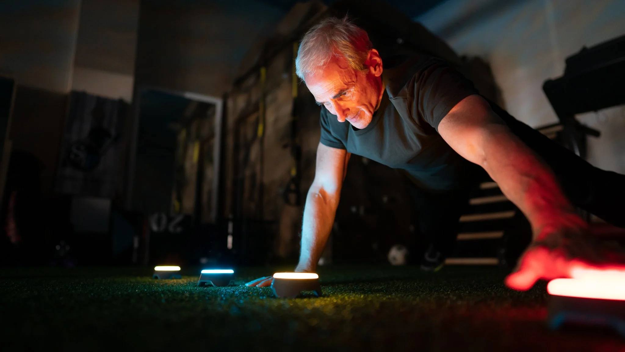 A man doing push-ups on a dark gym floor with illuminated workout equipment in front of him.