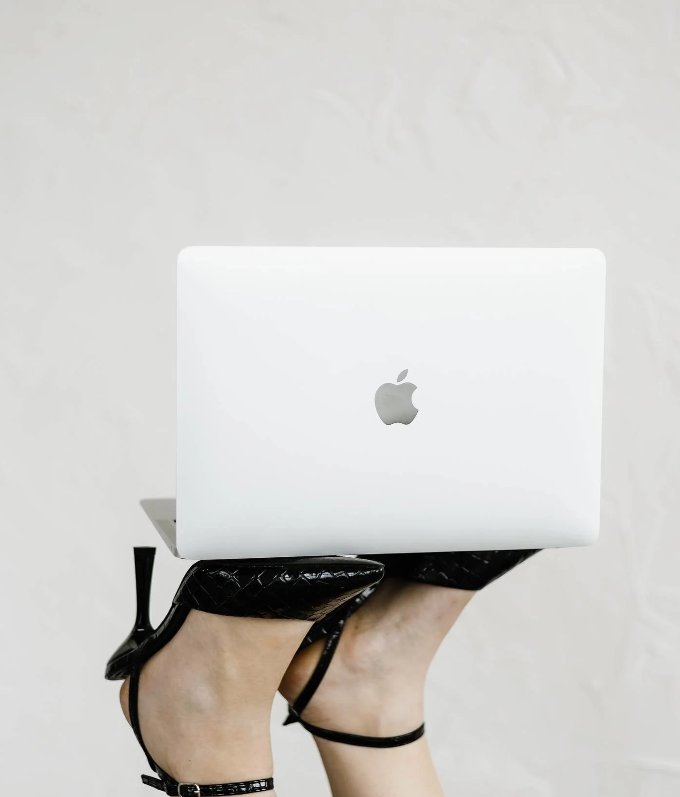 Person holding a white Apple MacBook laptop with a black strap around their wrist and black ankle strap, against a light background.