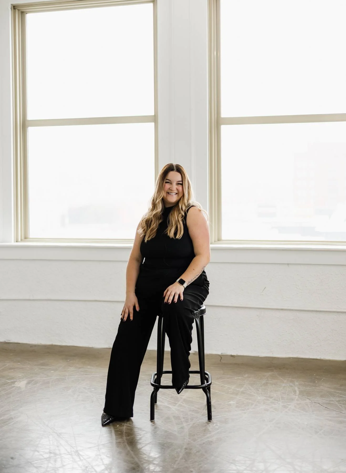 A woman with long wavy hair sitting on a black stool in front of two large windows with white frames, smiling and looking at the camera, wearing a black sleeveless top and black pants, with a smartwatch on her left wrist.