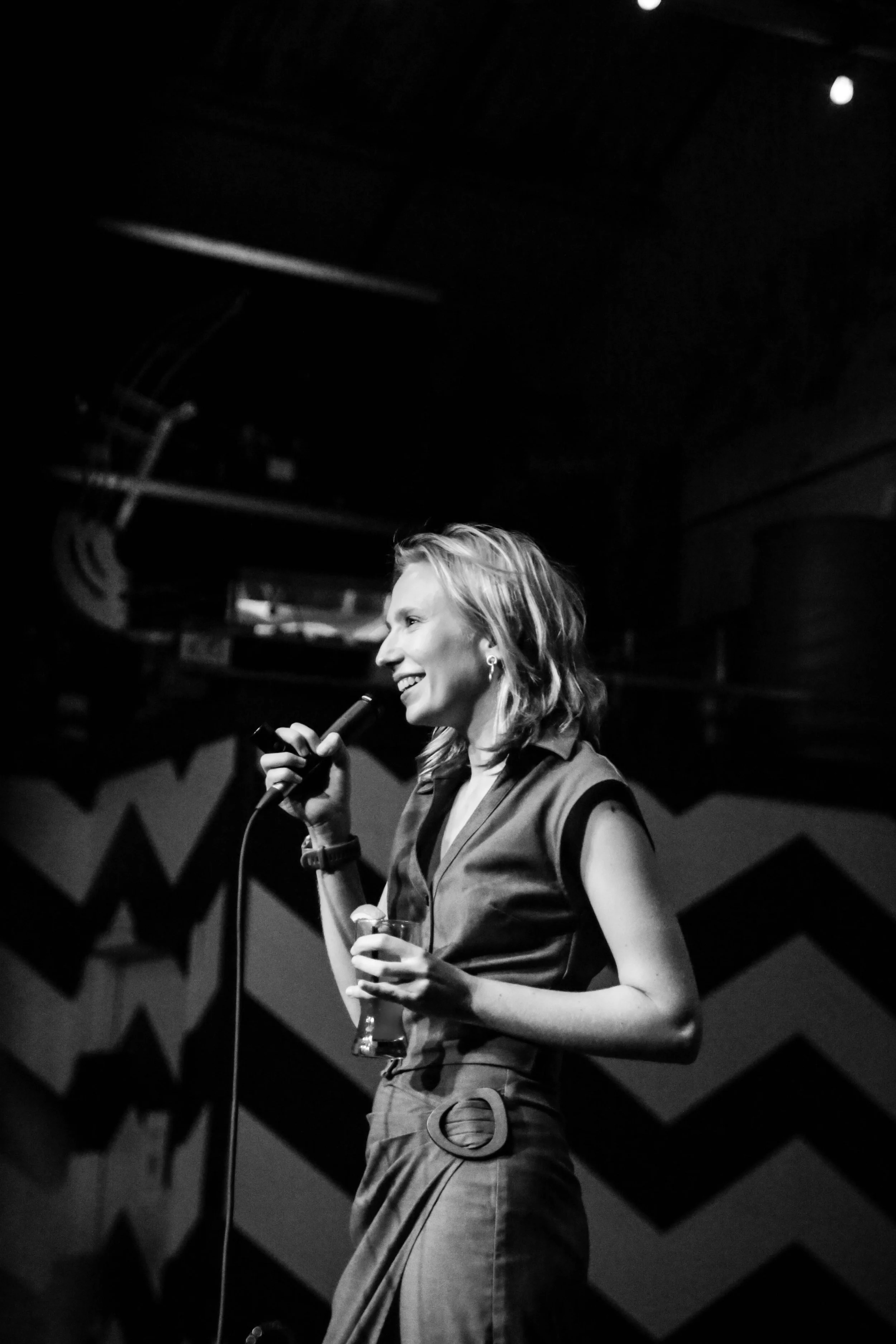Black and white photo of a woman with wavy hair, smiling, holding a microphone in one hand and a glass of drink in the other, standing on stage with a patterned background.