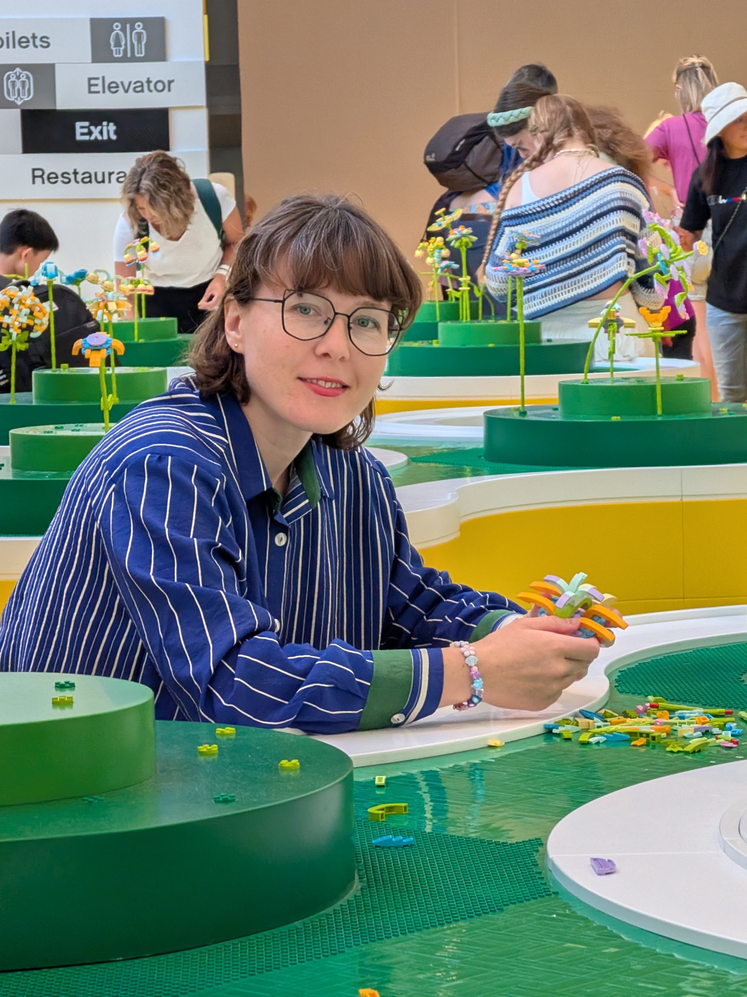Woman sitting at a table building with colorful LEGO bricks, with other people also working on LEGO projects in the background.