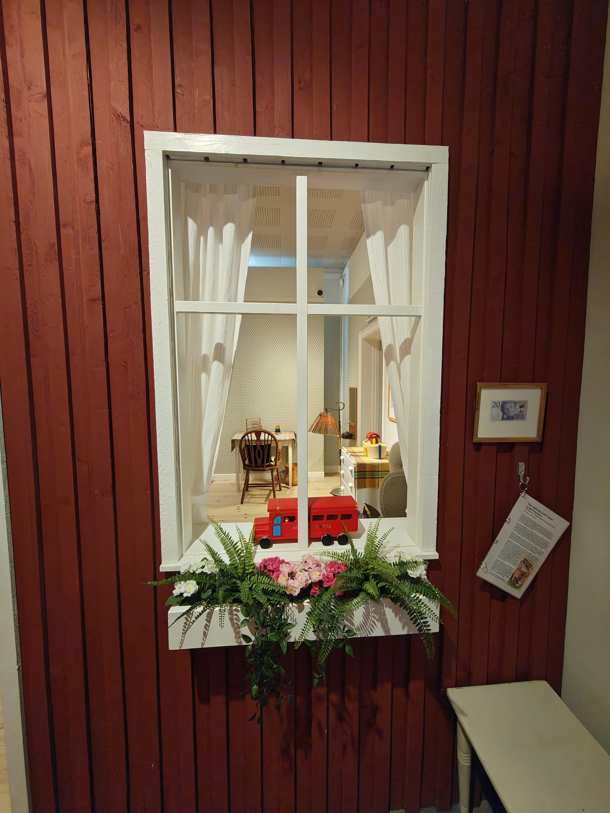 A view through a white-framed window with white curtains, showing a cozy room with a small table and chairs, a lamp, and various decorative items. The outside area has a brick wall painted red and a flower box with pink flowers and green plants.