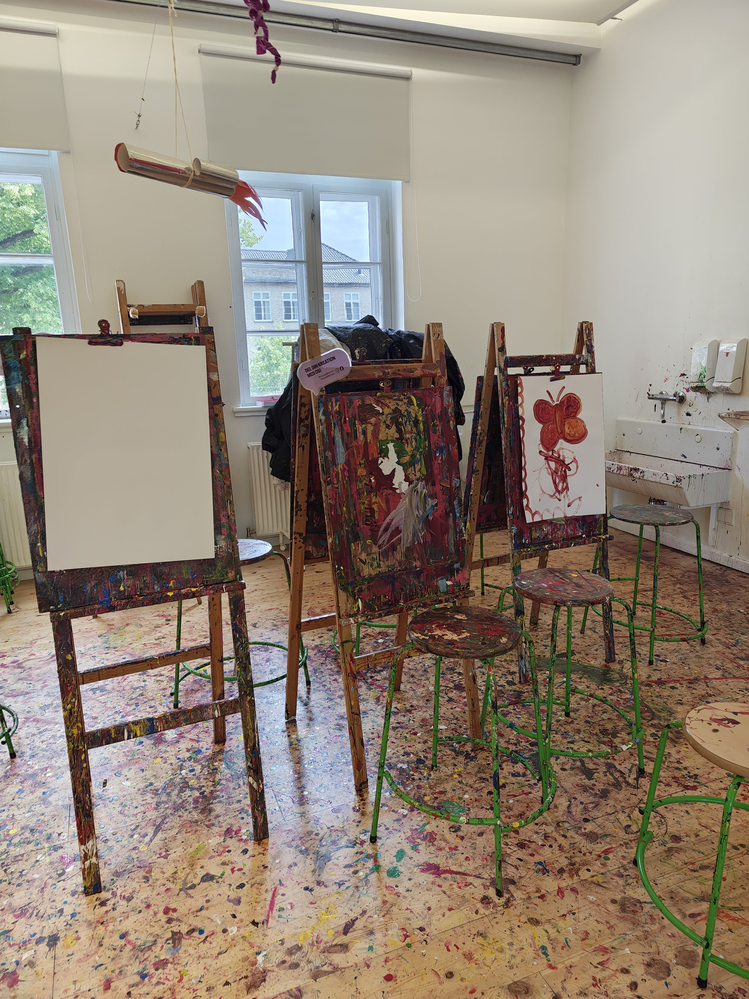An art classroom with canvases on easels, some with paintings, and a ceiling covered in colorful paint splatters. Natural light coming from windows, with paint-splattered floor and wall.