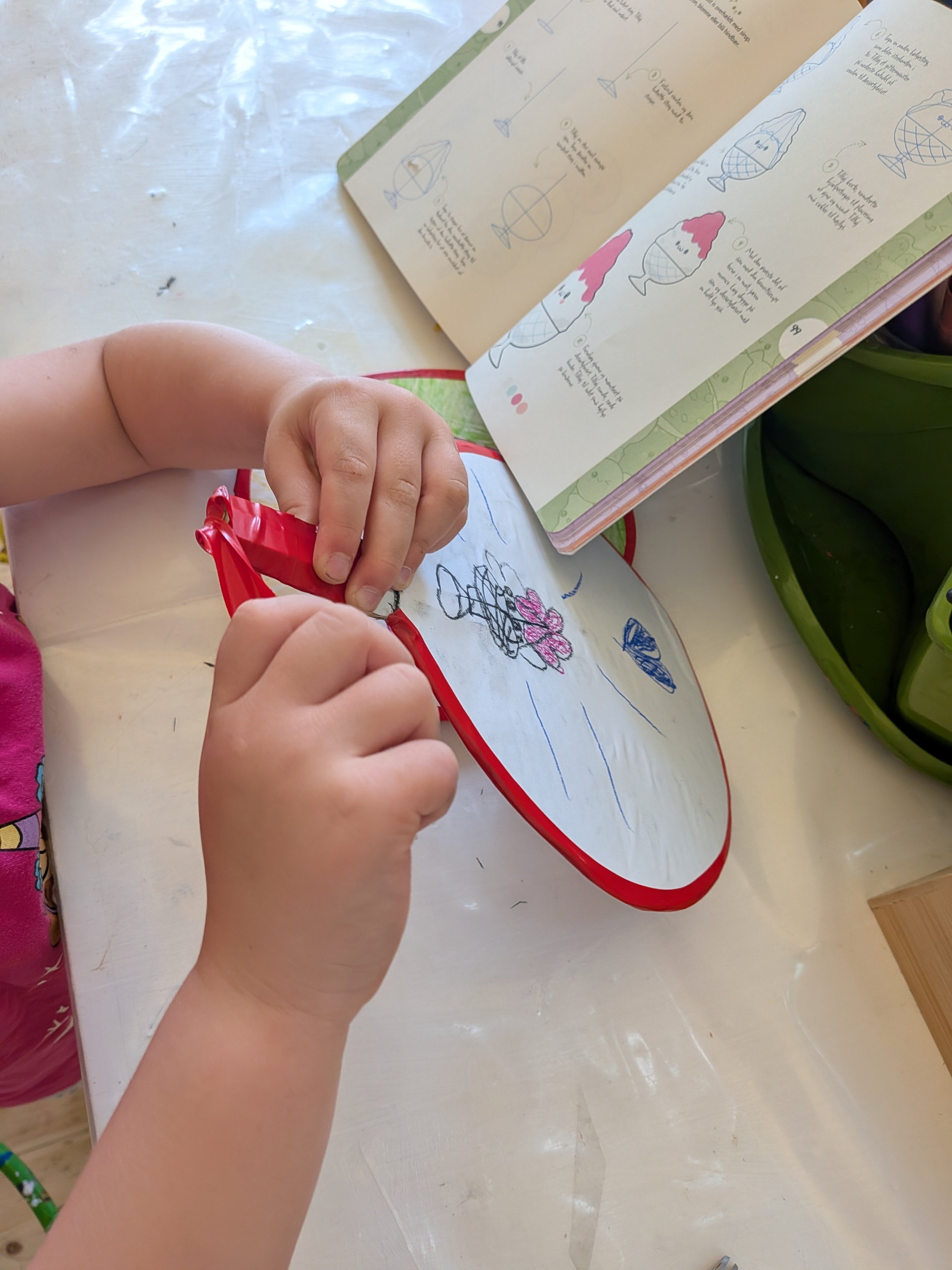 A child's hands holding a red and white cloth bag while drawing on a piece of paper with crayons, with an open instruction booklet with blue sketches of a fish and a water cage.