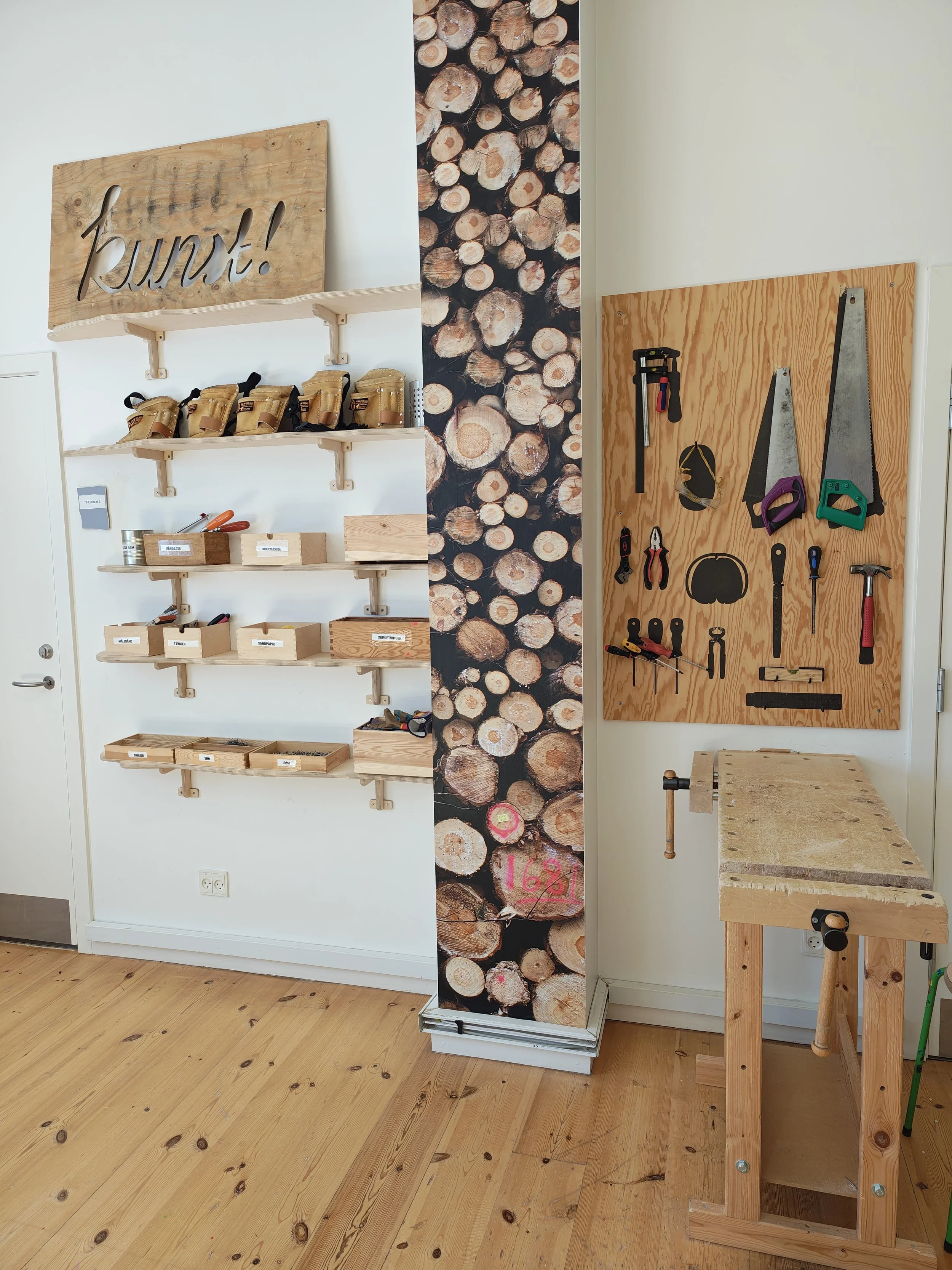A woodworking workspace with a wall-mounted wooden shelf holding several tool pouches, a large wooden board with various hand tools, a wooden workbench, and a decorative cardboard cutout of logs in the center.