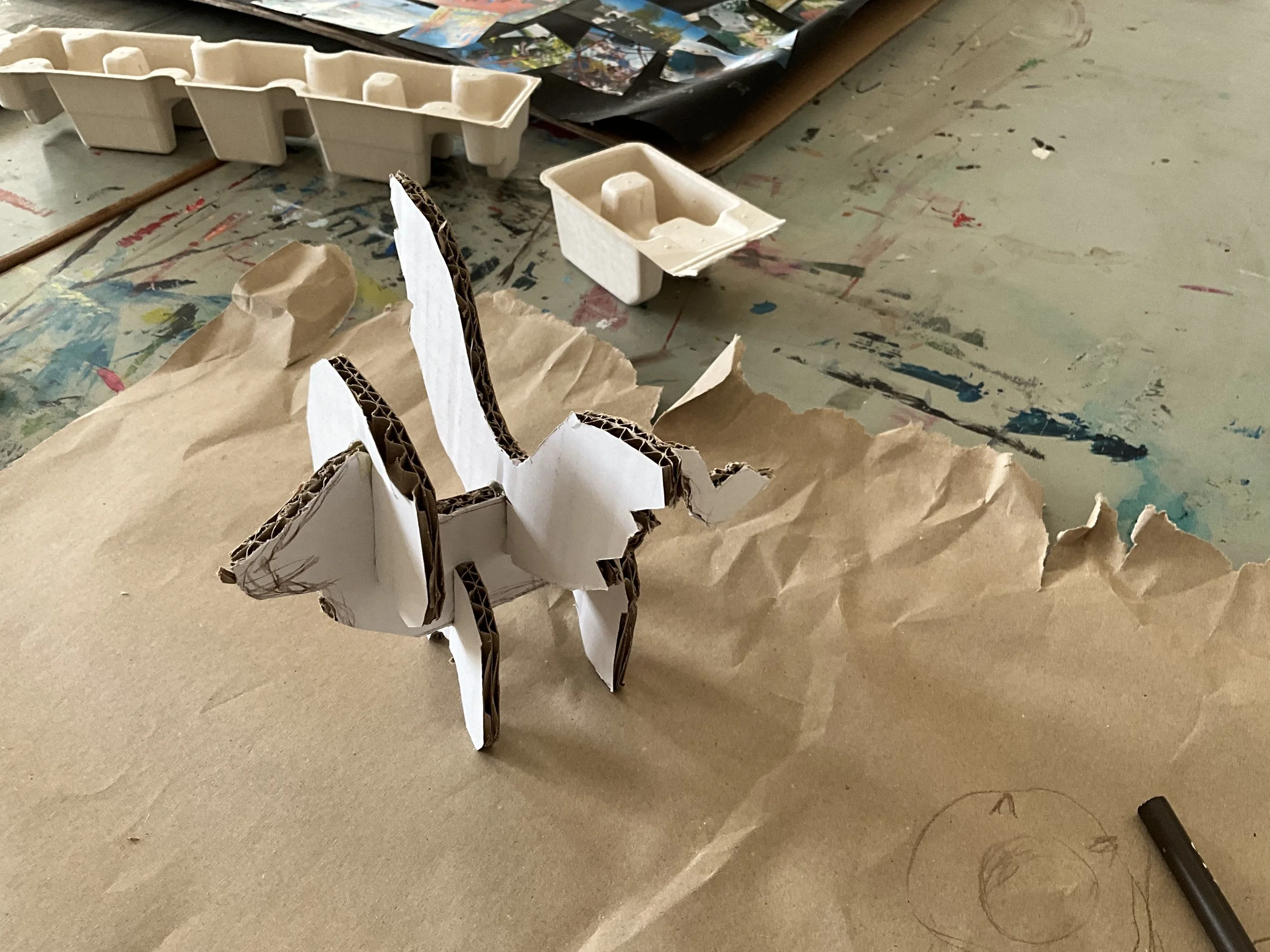 A messy table with crumpled brown paper, a small white cardboard model of a horse, and various art supplies including torn paper and paints.
