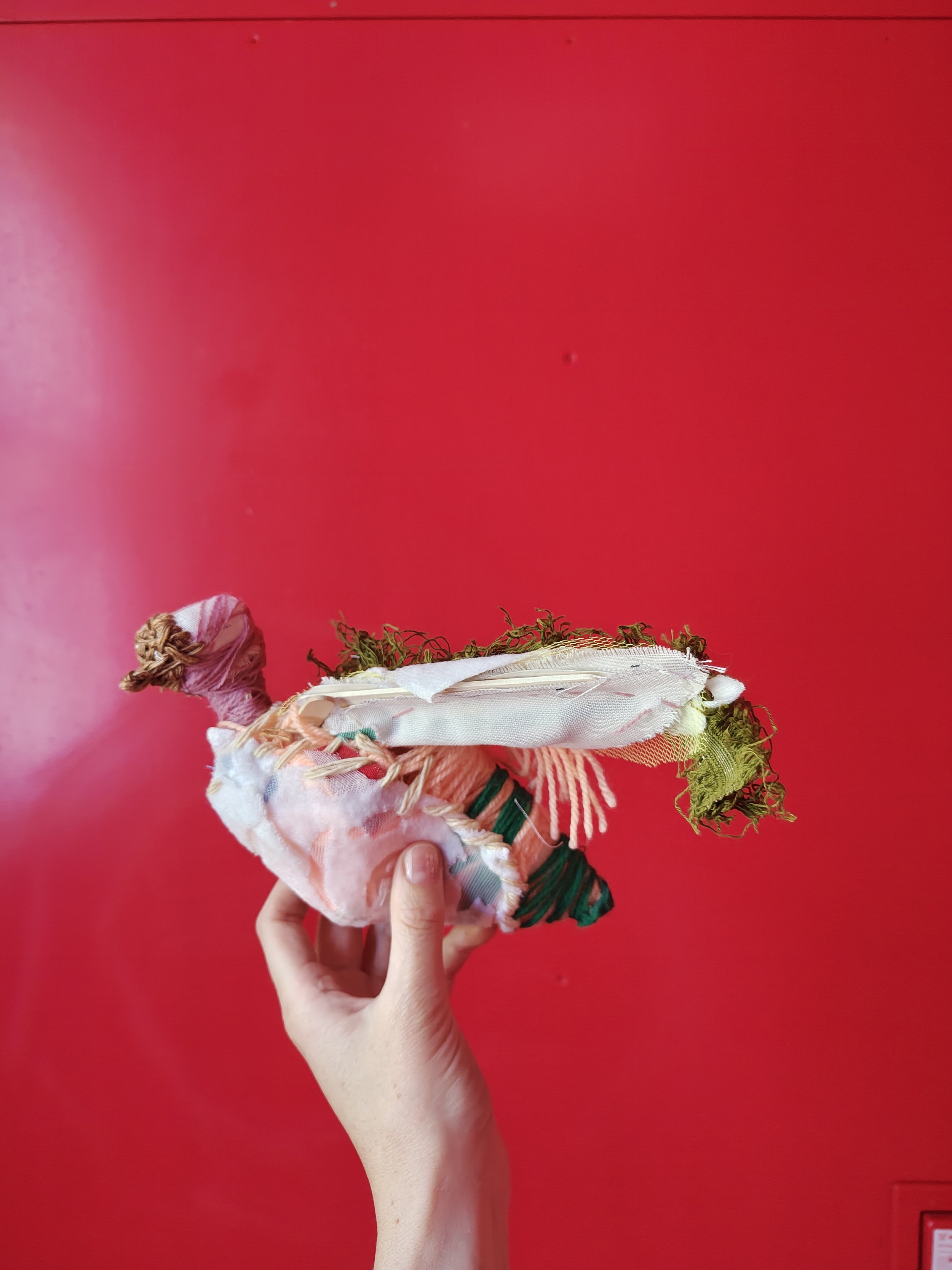 Hand holding a fabric and yarn sculpture of a unicorn against a bright red wall.