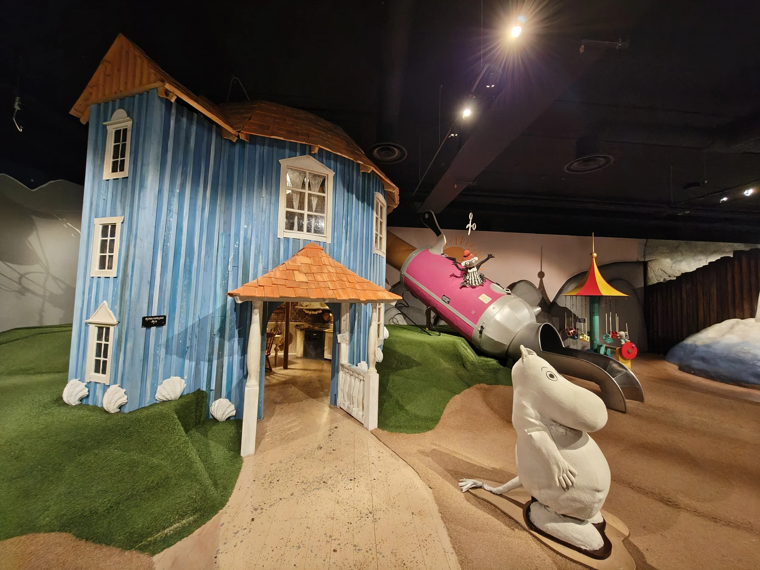 Indoor play area featuring a large blue wooden house with white window frames, a small porch, and a red shingle roof. There are artificial grass patches, a pink helium balloon sculpture of a character with arms raised, and a white swan-shaped ride. The ceiling has bright lights and dark panels.