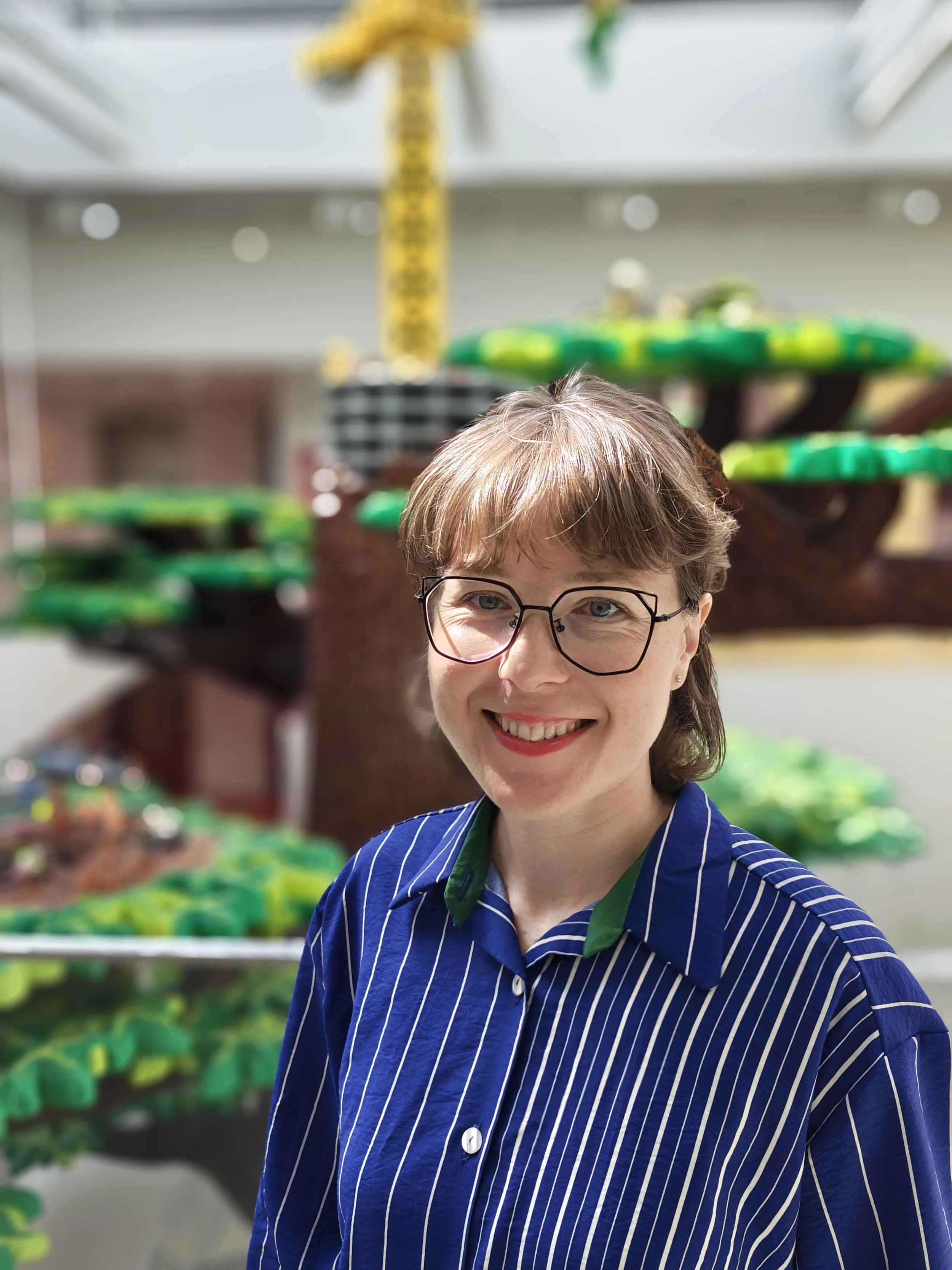 A smiling woman with short brown hair and glasses wearing a blue striped shirt, standing outdoors in front of colorful climbing toys.