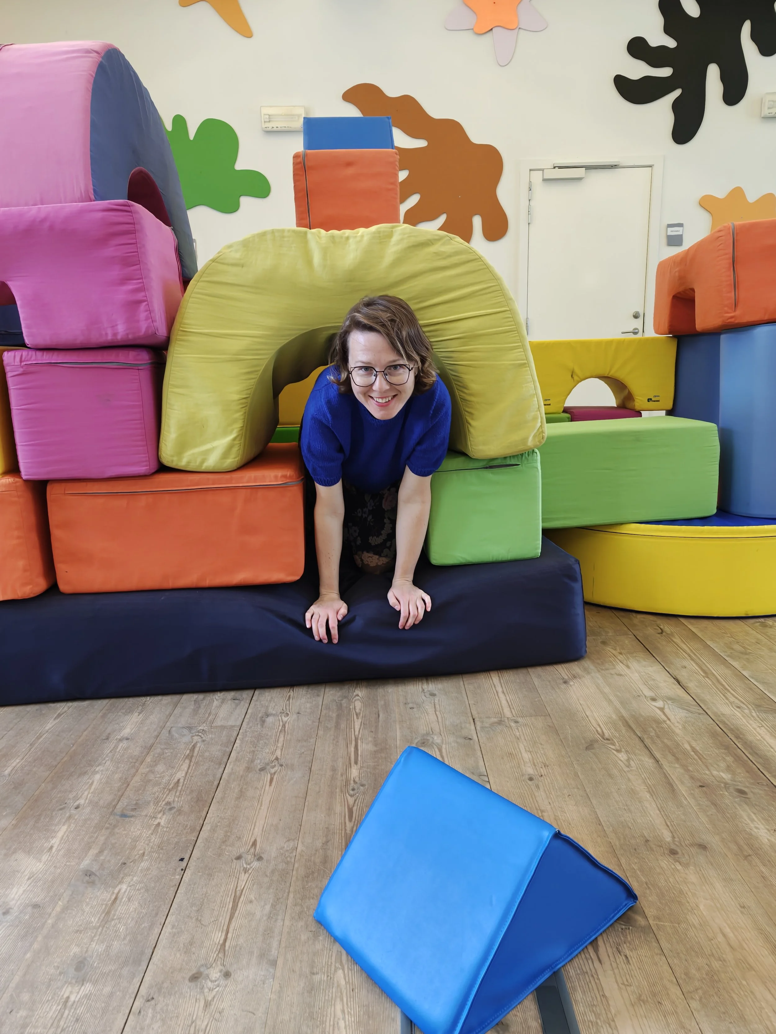 A woman with glasses is crawling through a soft play structure with colorful foam blocks in a play area. There is a blue foam block on the floor in the foreground. The background features foam blocks in pink, yellow, green, orange, blue, and black, arranged in various shapes and sizes.