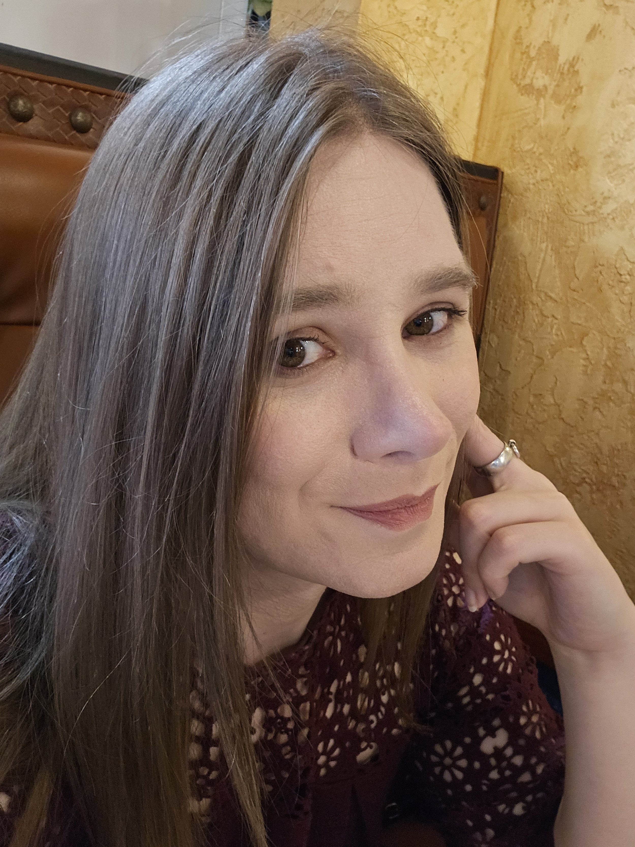 A close-up of a woman with long, blondish-brown hair, wearing a maroon, crochet pattern top, and a silver ring on her finger, sitting in a room with a yellow textured wall and a wooden headboard.