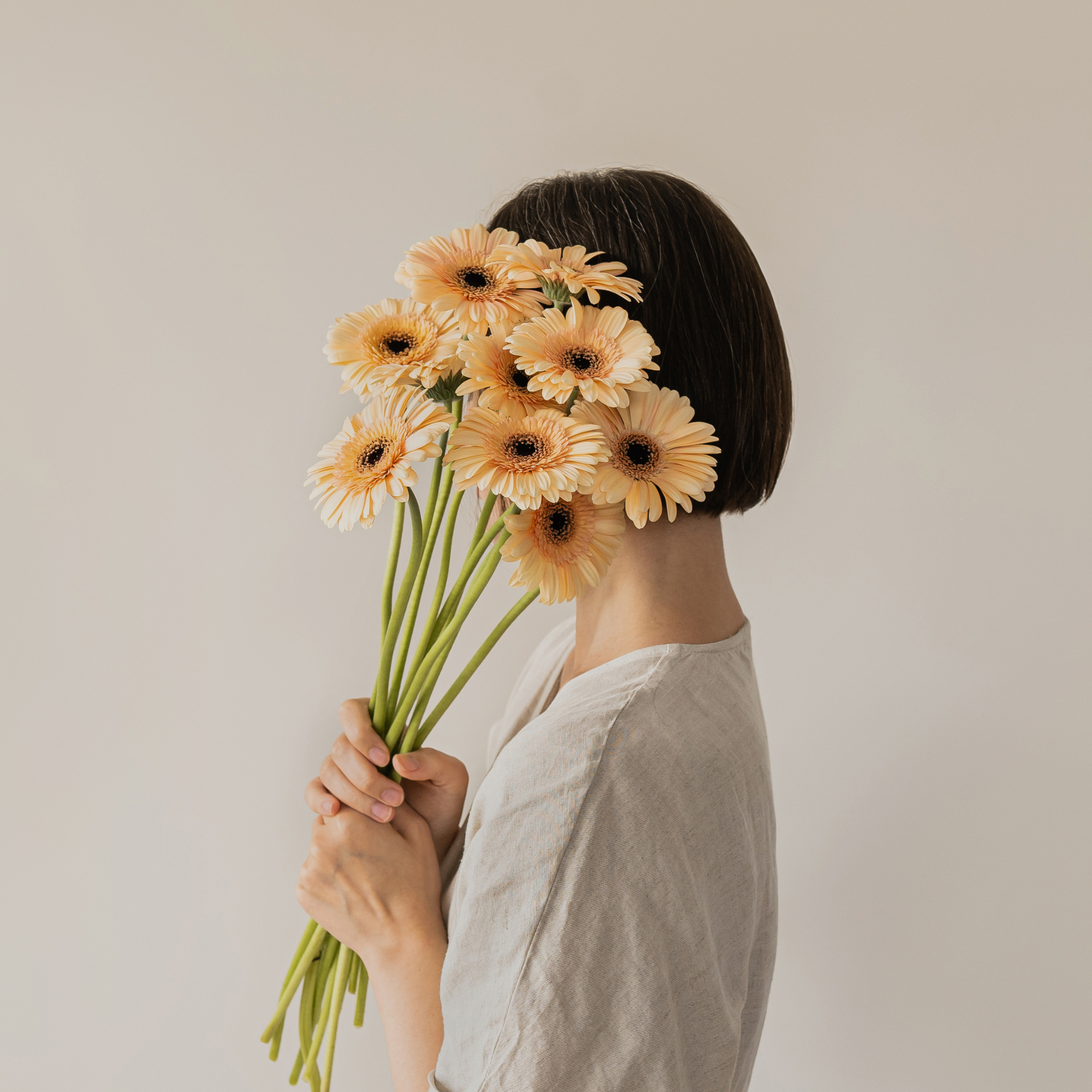 A woman with short dark hair wearing a beige shirt, holding a large bouquet of peach-colored daisies with black centers in front of her face, standing against a plain light gray background.
